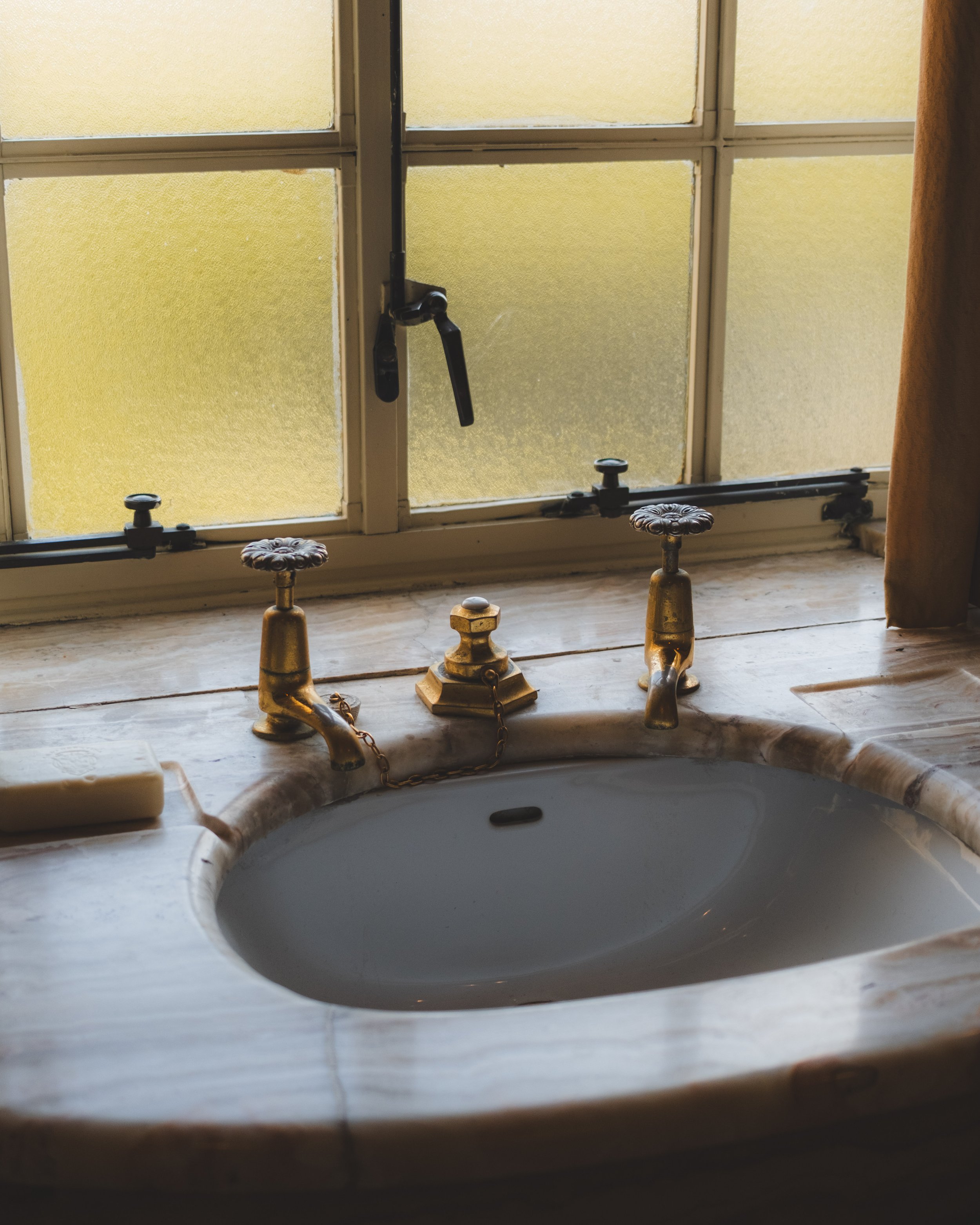 A bathroom window with textured yellow glass panes and a brass-colored faucet with chains, a marble sink edge, and a white bathtub below.