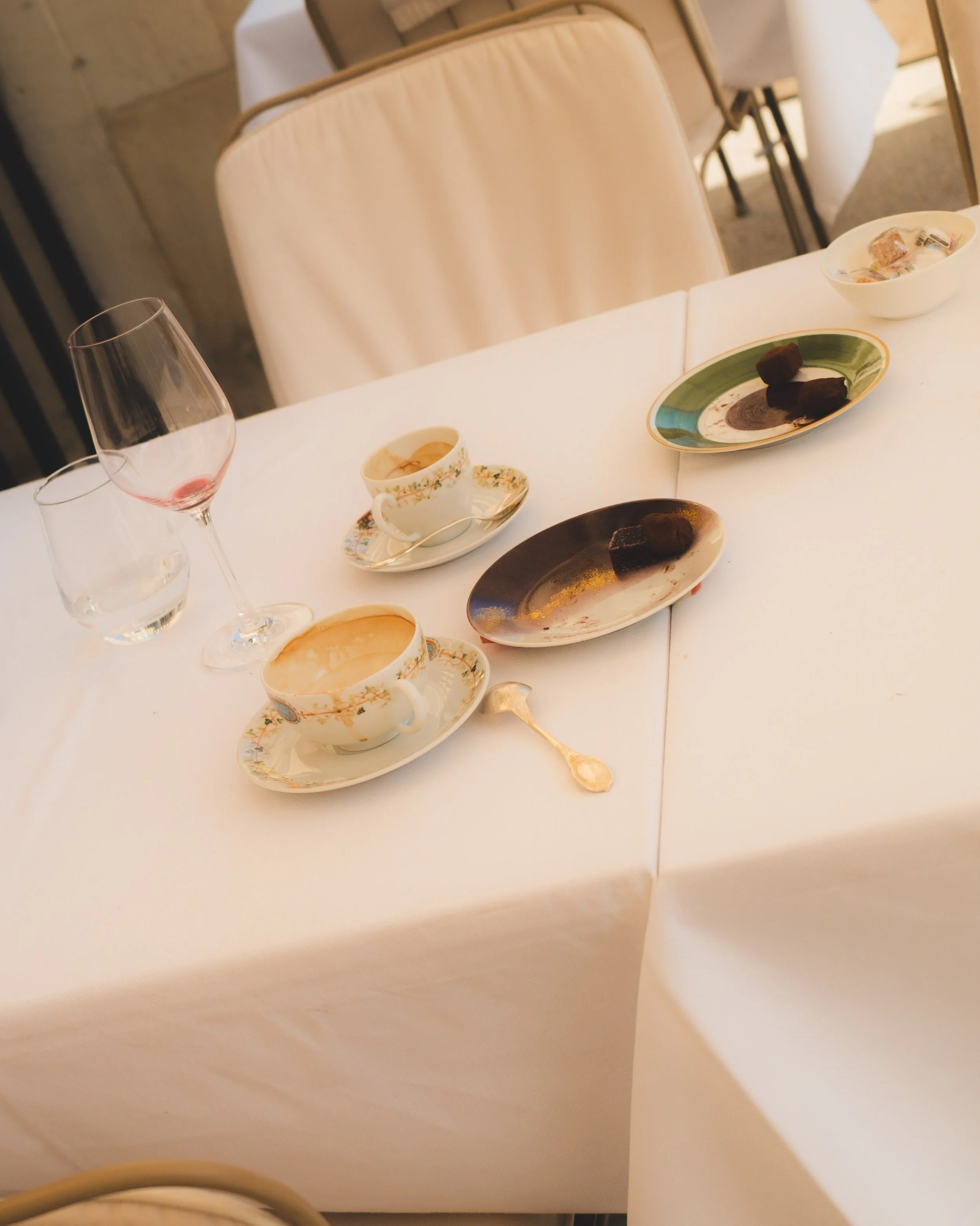 A table set with a largely white tablecloth, various cups, glasses, and plates with desserts and candies, and a chair with a cream-colored cover is seen in a dining area with a wooden wall in the background.
