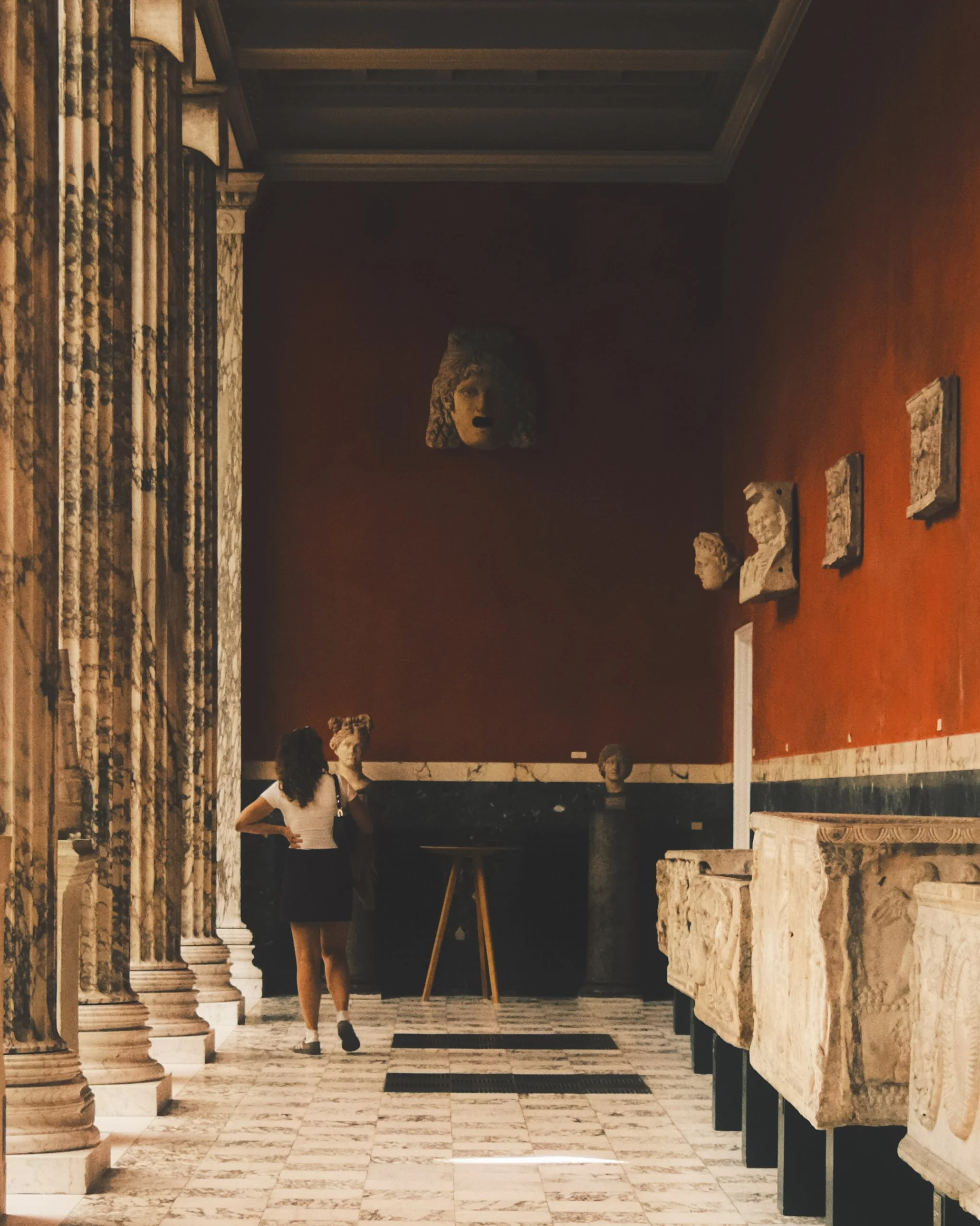 Interior of a museum with classical sculptures and columns. A woman stands in the background, looking at the exhibits. The room has stone flooring and a warm-colored wall decorated with sculptures on plaques.