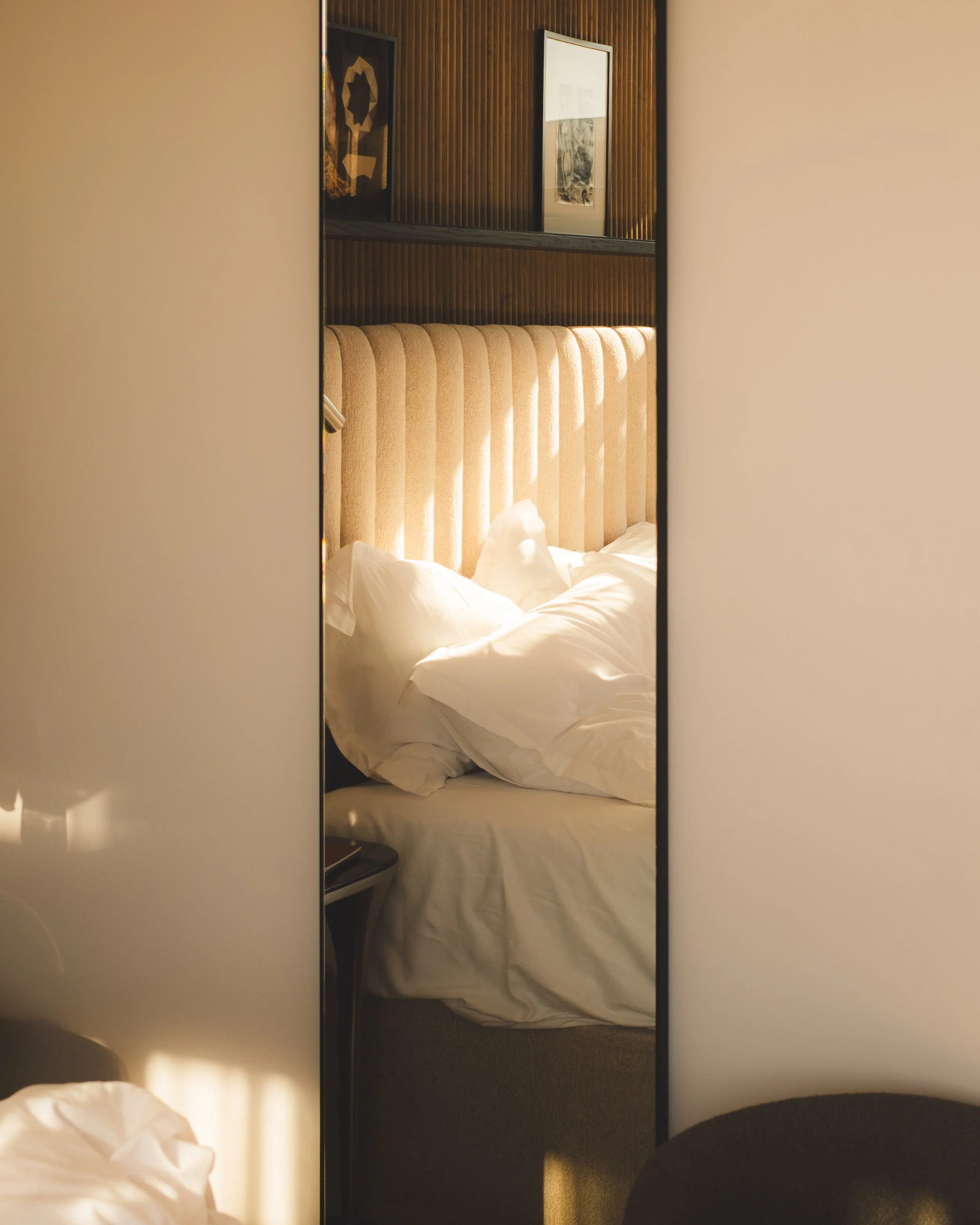Photo of a hotel room interior as seen through a mirror, showing a bed with white sheets and pillows, a wooden headboard, framed artworks on a wood-paneled wall, and a nightstand.