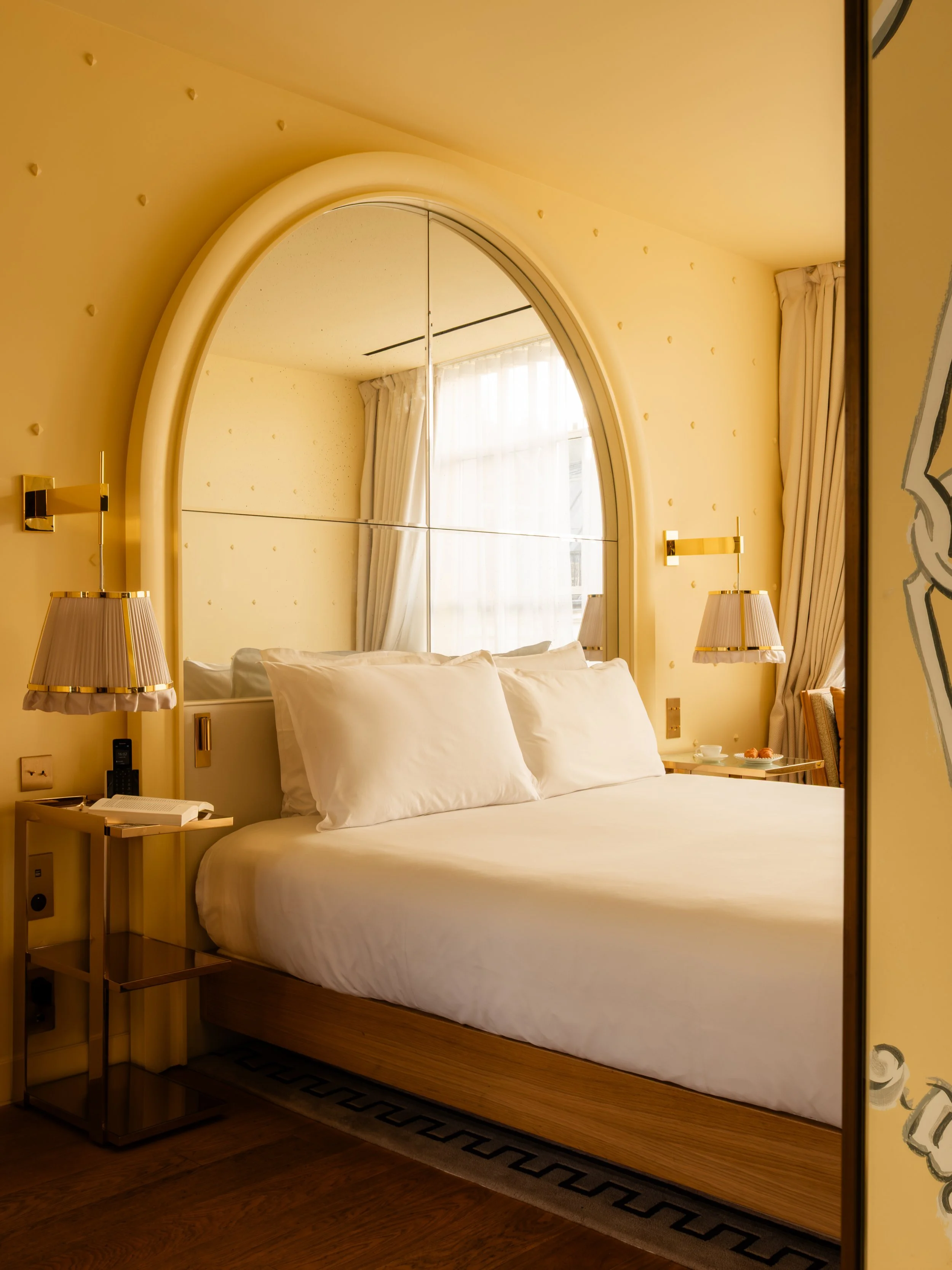 A hotel room with a bed, white linens, large pillows, and a mirror headboard. Two bedside tables with lamps and curtains near a window. Lionel Chu - Hotel & Travel Photography