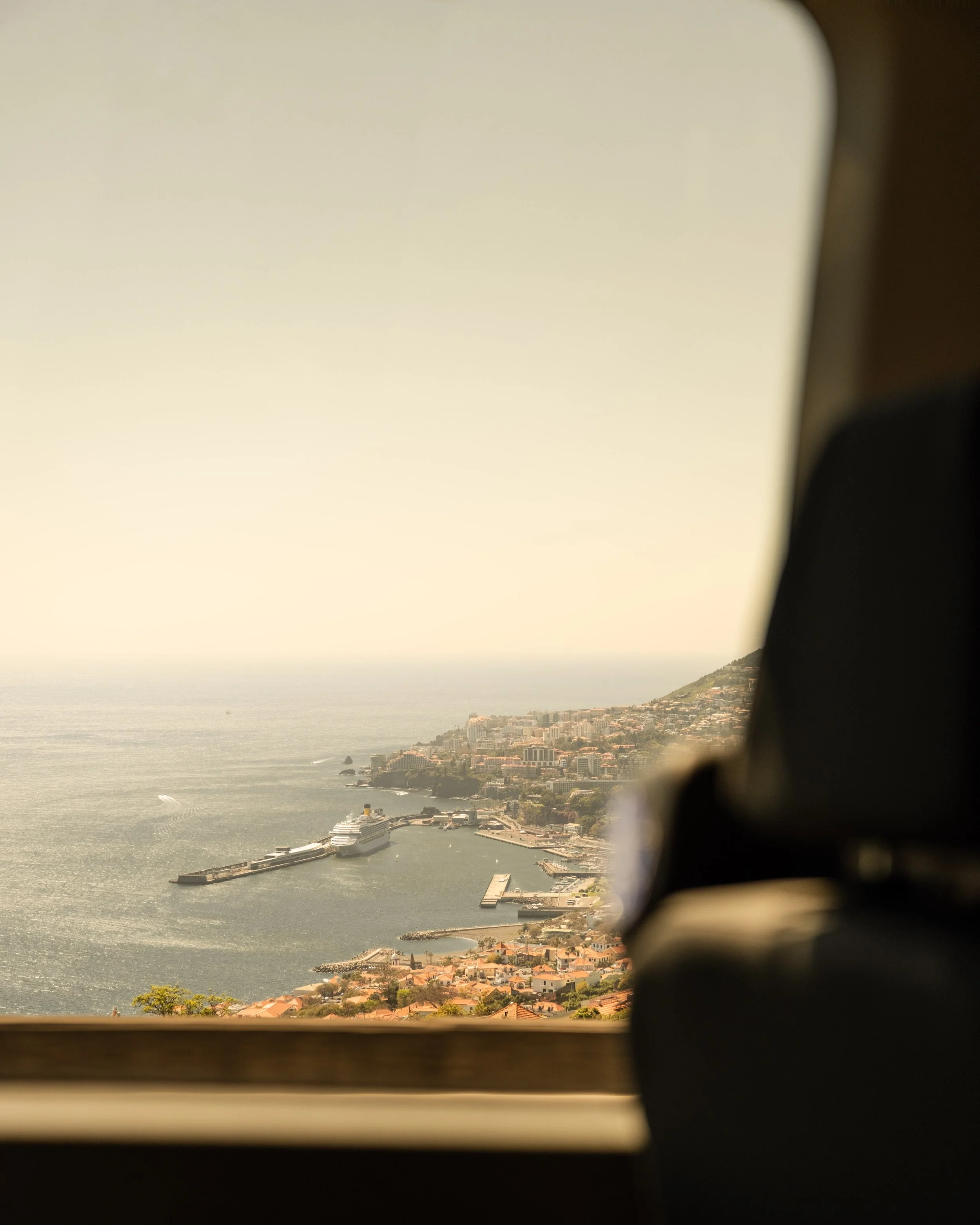 An aerial view from a car window of a coastal city with a cruise ship docked at the pier, surrounded by buildings, hills, and the ocean under a hazy sky.