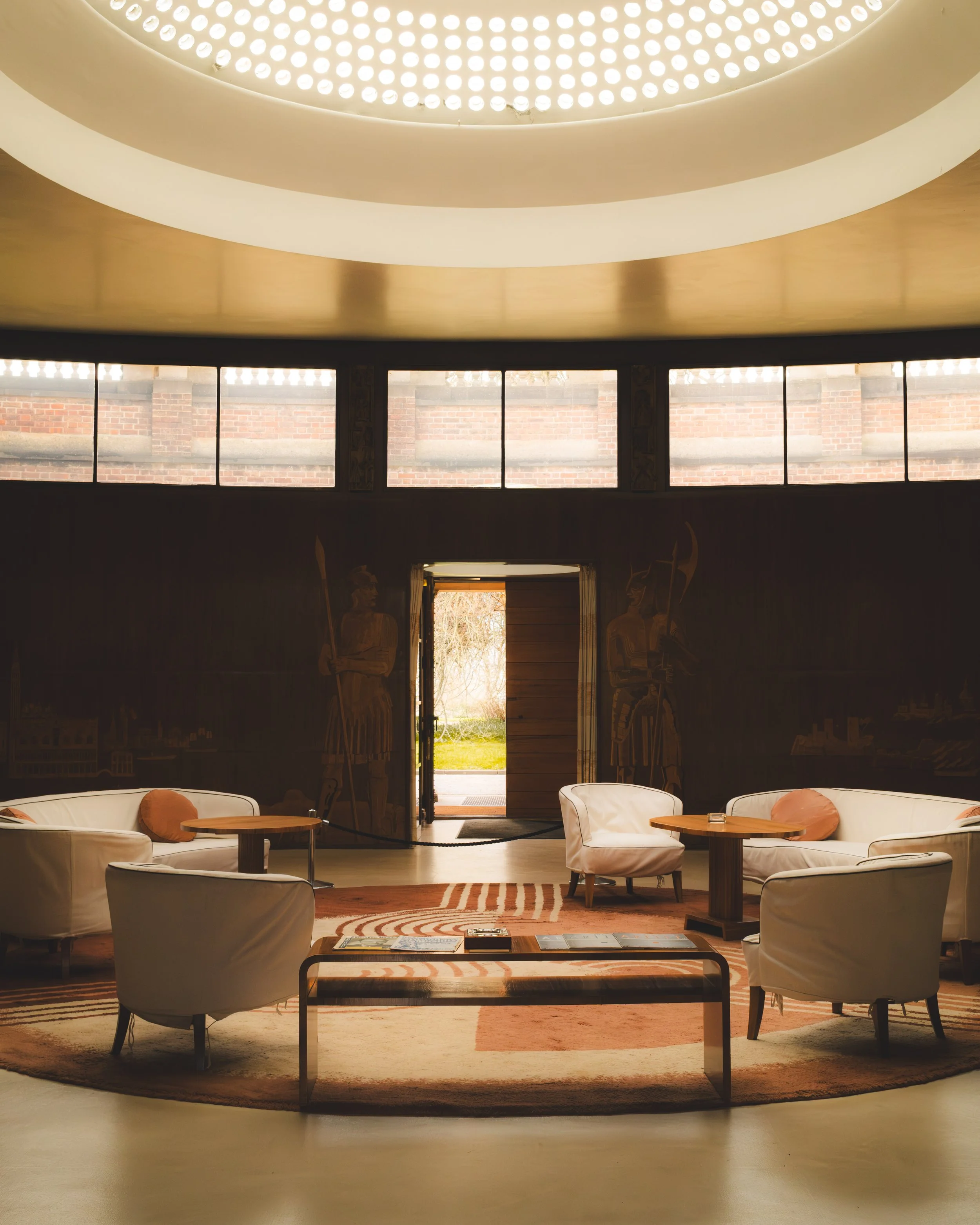 Inside a room with large windows and a high ceiling with round recessed lighting. There are white upholstered chairs and small wooden tables, with a patterned rug on the floor. A door leads outside to a grassy yard.