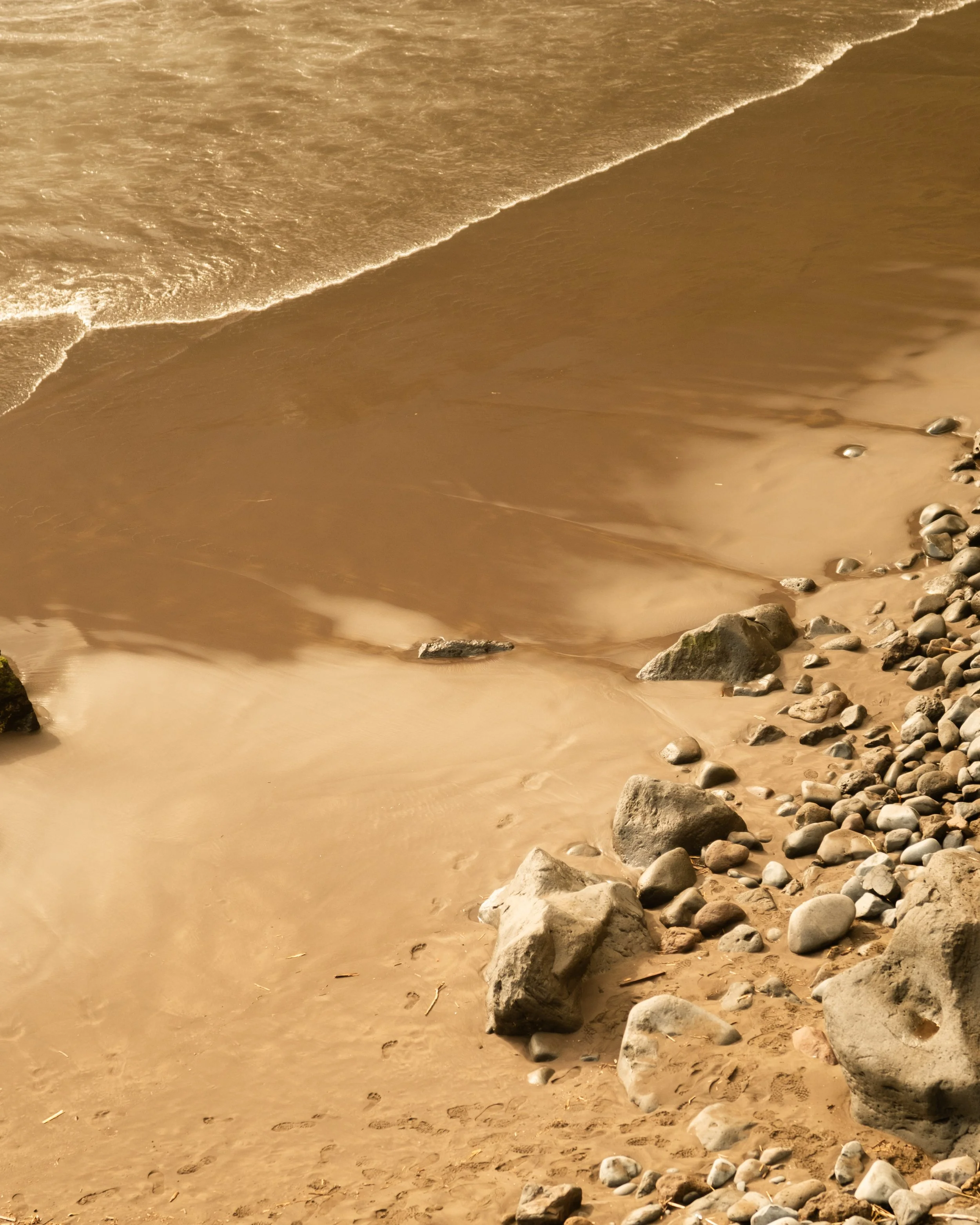 A sandy beach with rocks along the shoreline and gentle waves lapping onto the sand in an orange or sepia tone.