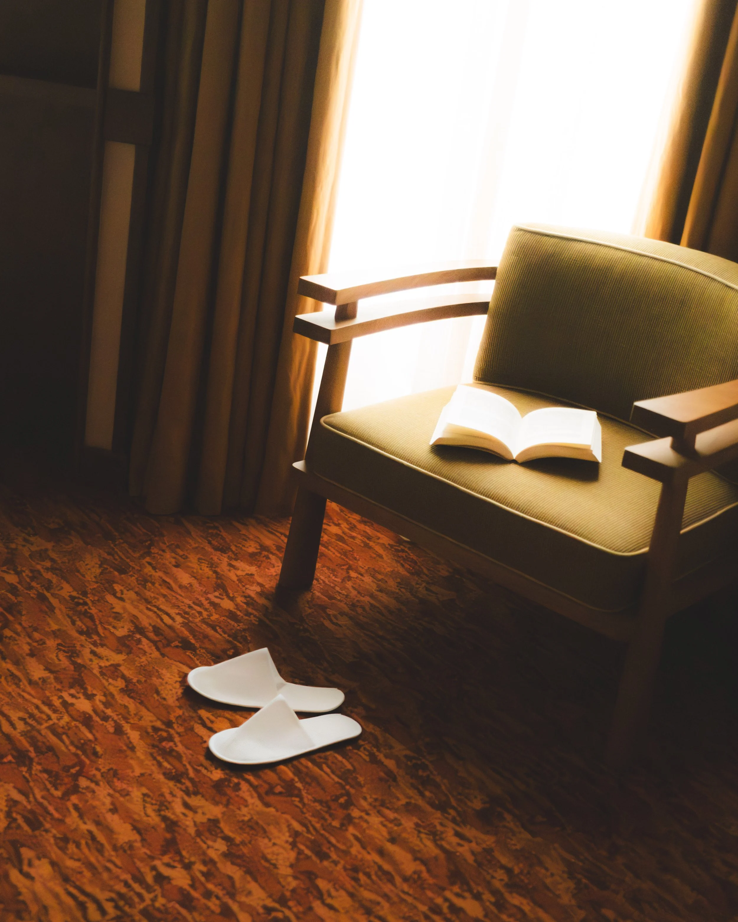 A green upholstered chair with armrests and a book open on its seat, in front of a window with white sheer curtains and heavy brown drapes, on a wooden floor with a patterned carpet.