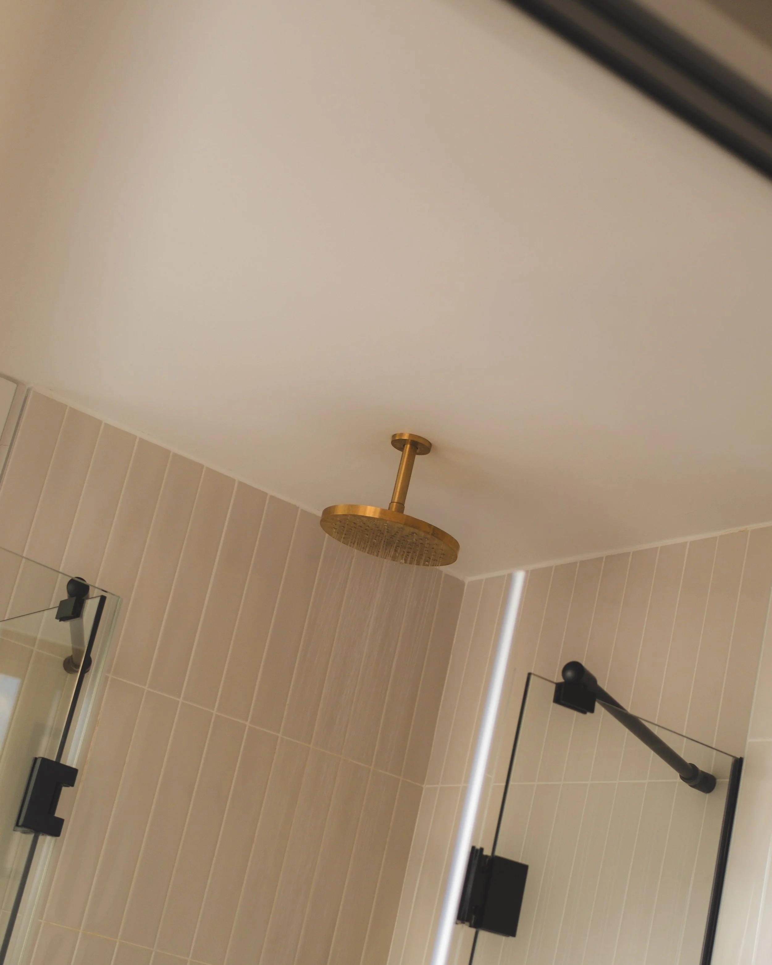 A bathroom mirror reflecting a showerhead and tiled wall, with a black and brass shower fixture and a portion of a glass shower door.
