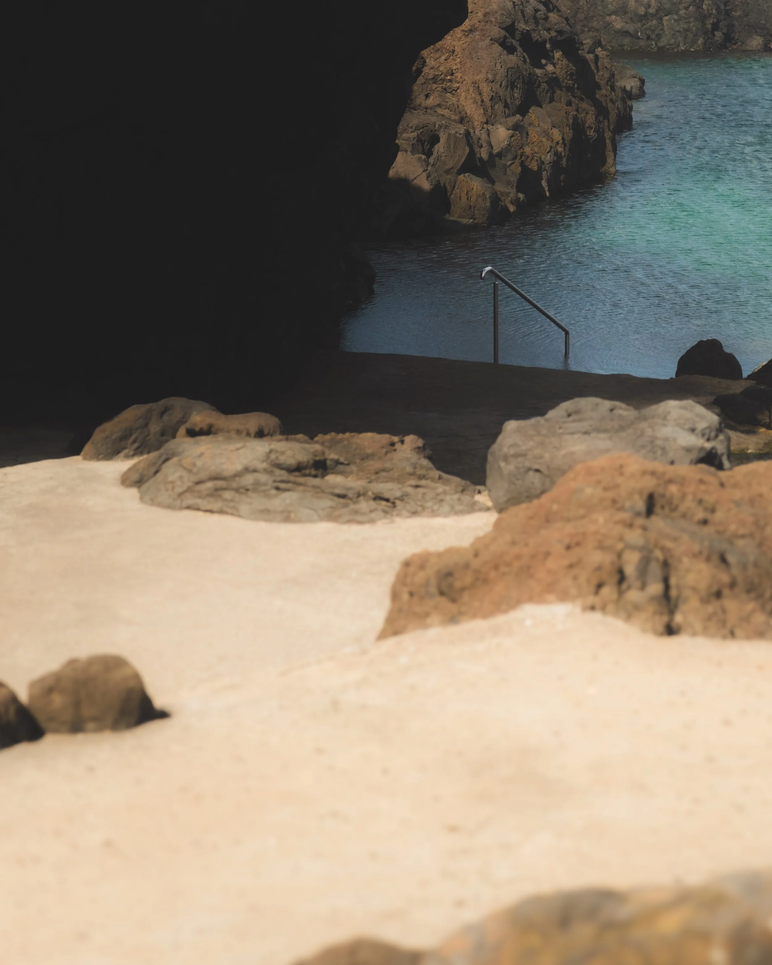 View from a beach looking out towards a rocky shoreline with water, a metal railing extends into the water, and rocks are scattered in the foreground.