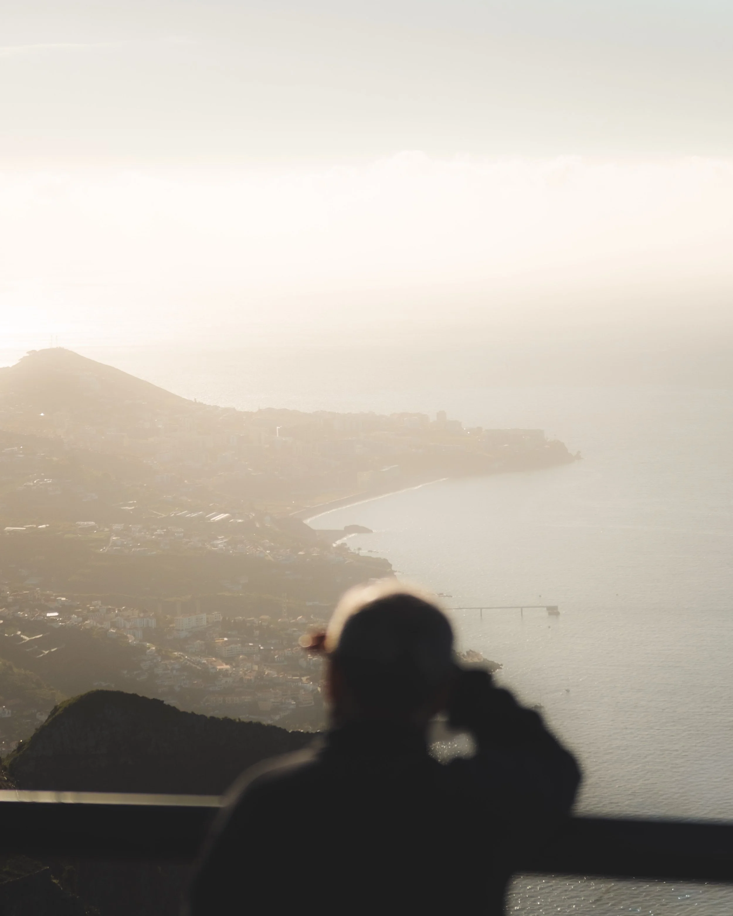 A person is sitting on a ledge facing a scenic view of hills and a city, with a hazy sunset or sunrise illuminating the landscape.