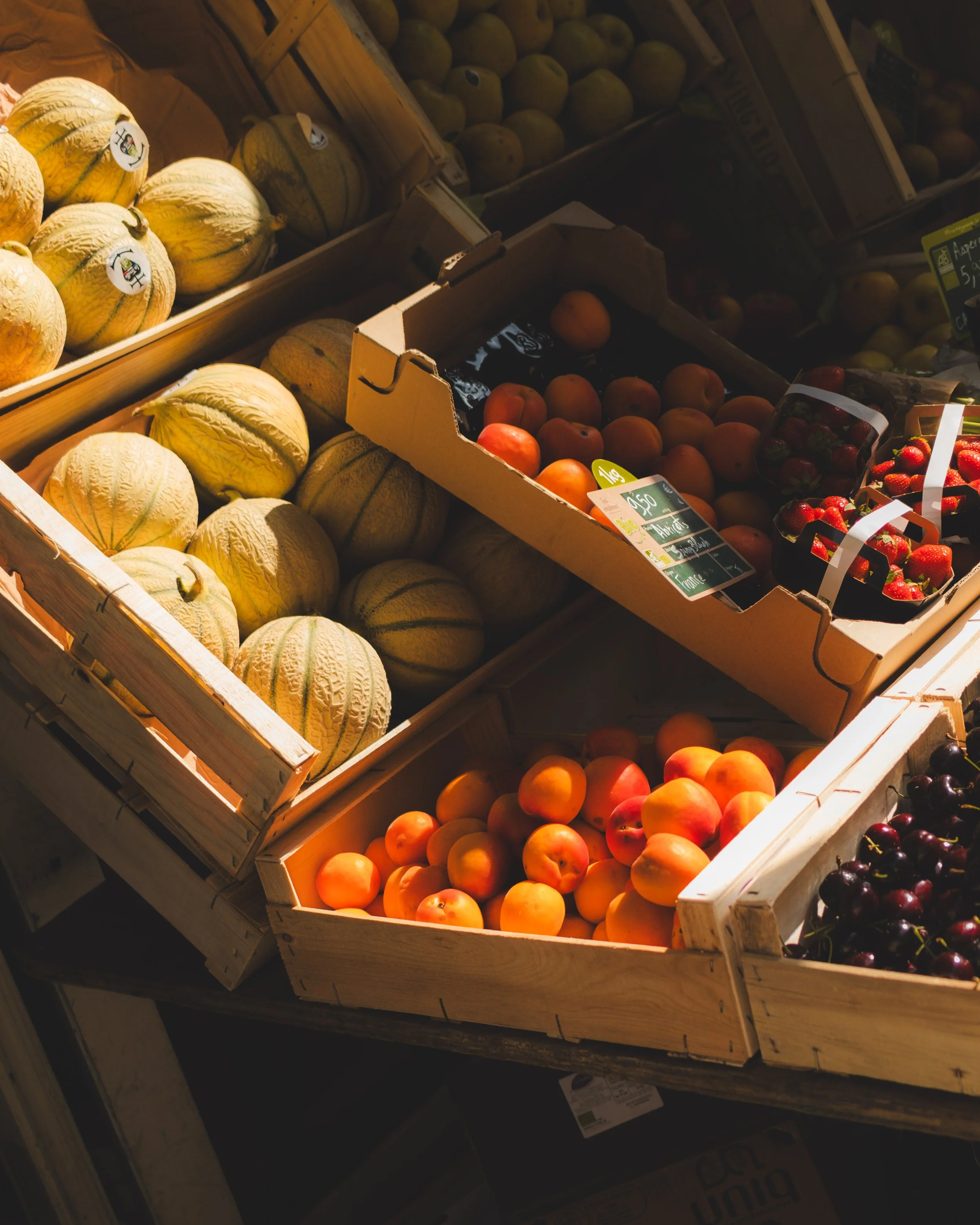 Wooden crates filled with cantaloupe melons, peaches, and strawberries at a grocery store produce section.