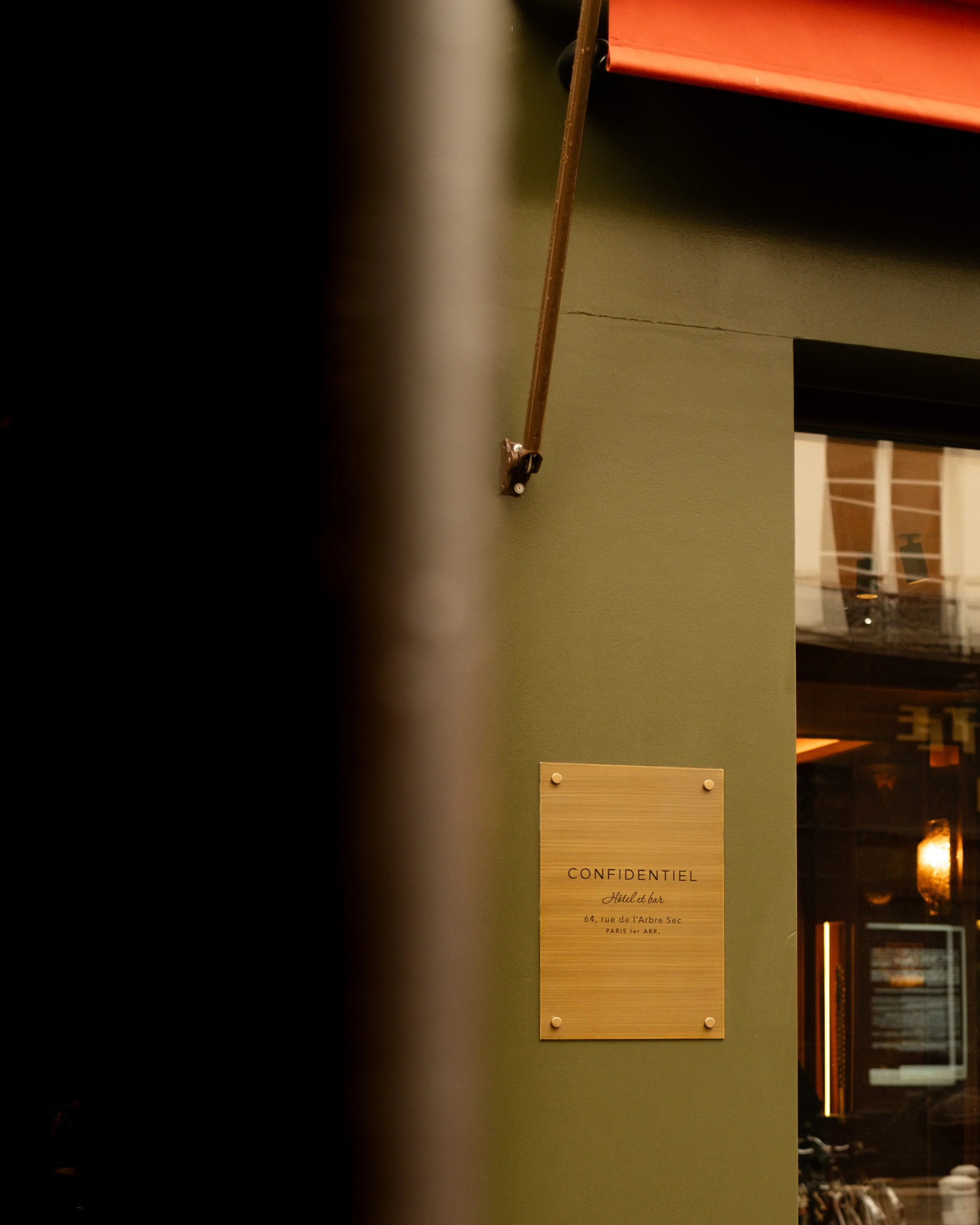 A partially obscured view of a green wall with a gold-colored plaque that reads "Confidentiel, Hotel & bar, 64, rue de l'Arbre Sec, Paris 1er Arr." to the right, with a window showing an interior with warm lighting. 