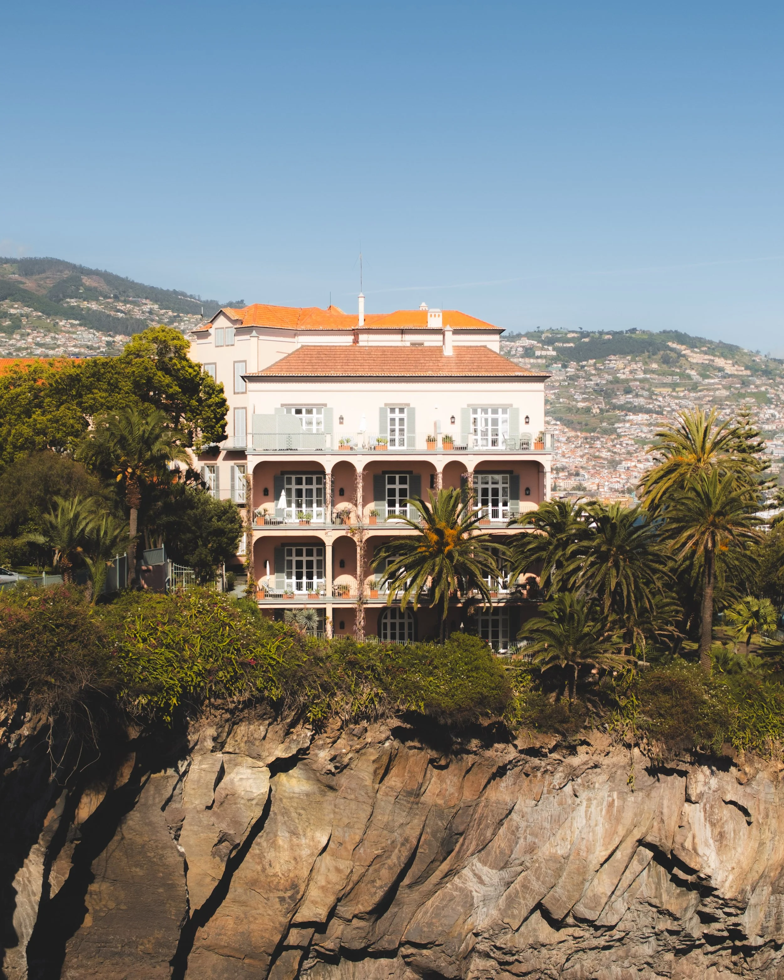 A view of a the Reid's Palace hotel with orange-tile roof and white walls, partly obscured by lush green trees, with a hillside cityscape in the background under a clear blue sky. The luxury hotel is located above a rocky cliff.