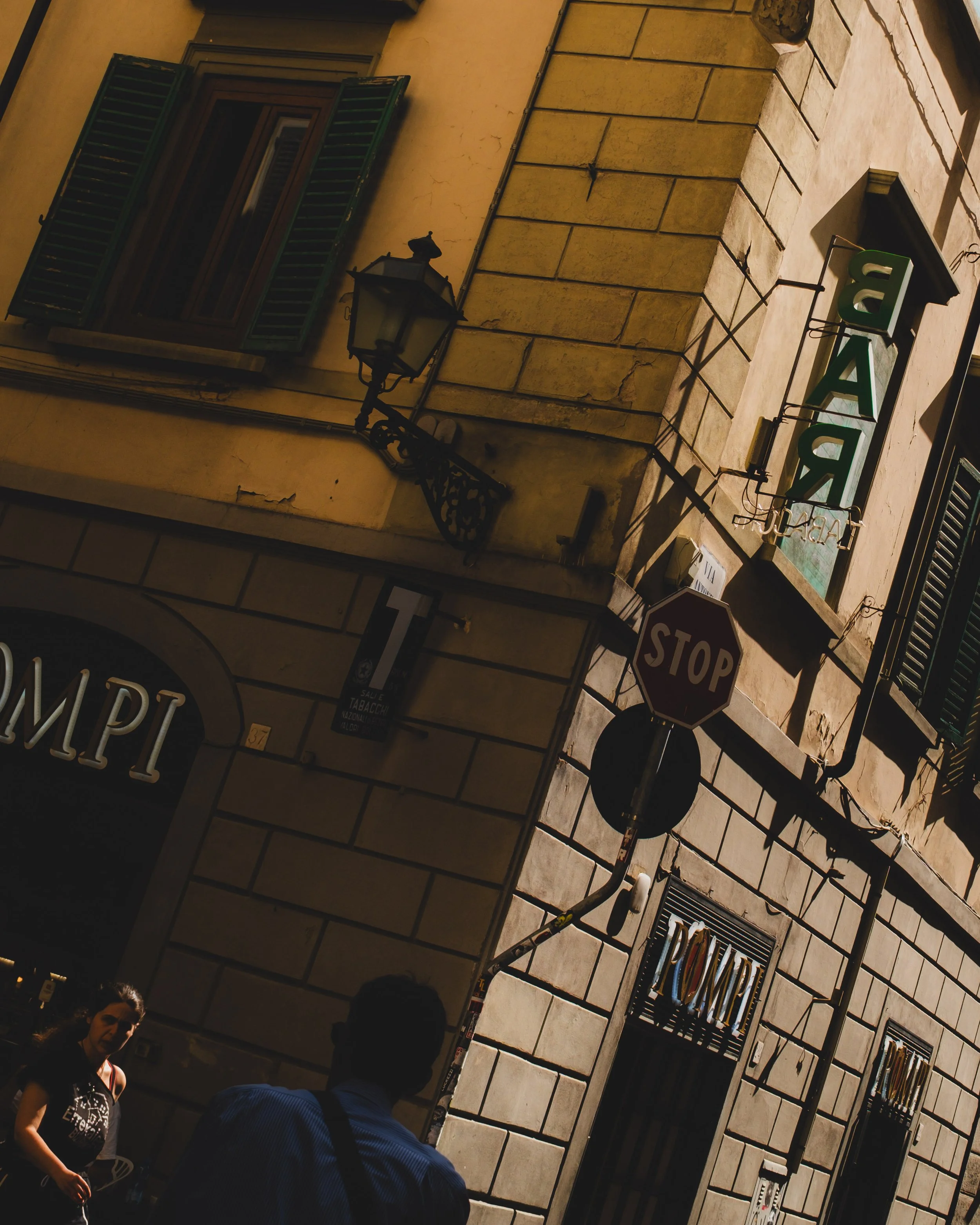 A street corner in Italy with a building featuring green shutters, a street lamp, a stop sign, and a neon sign that says 'BAR'. There are two people in the foreground, one woman and one man, near a shop with a sign written in Italian. Lionel Chu - Ho