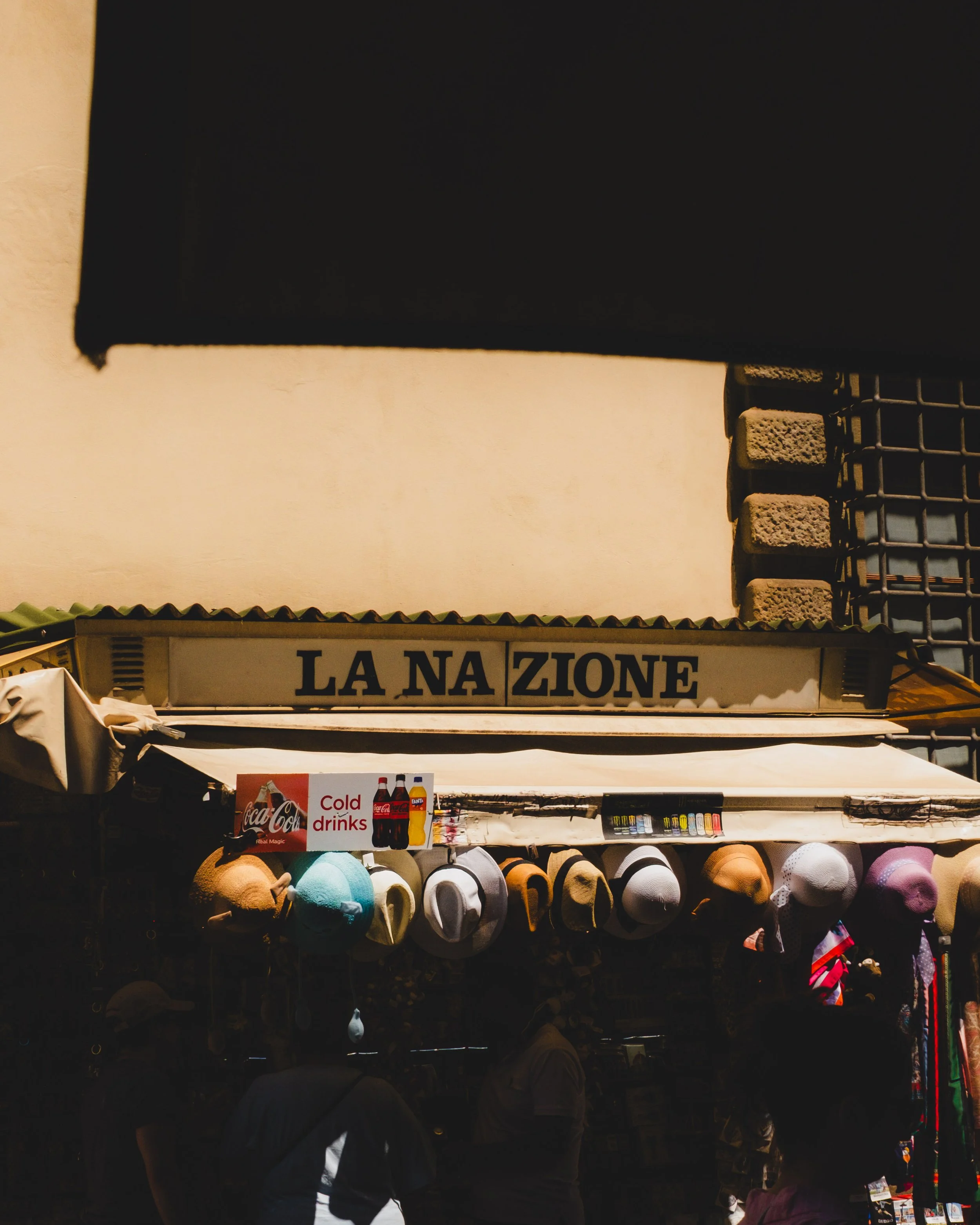 A street stall with hats and signs, including one for cold drinks, located under a sign that reads 'LA NAZIONE' in an outdoor market scene. Lionel Chu - Hotel & Travel Photographer