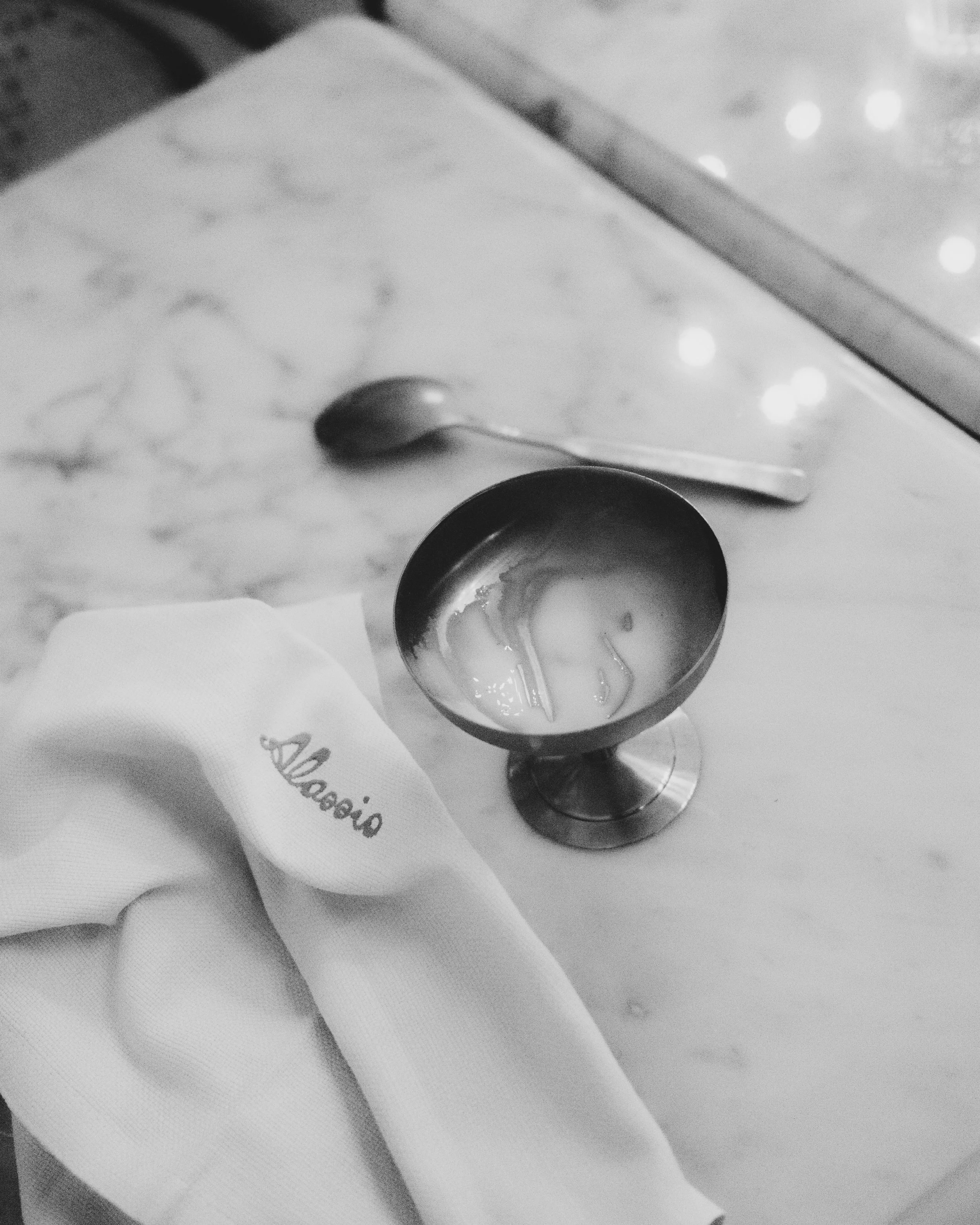 A close-up black-and-white photo of a metal spoon and a metallic goblet on a table with a napkin.