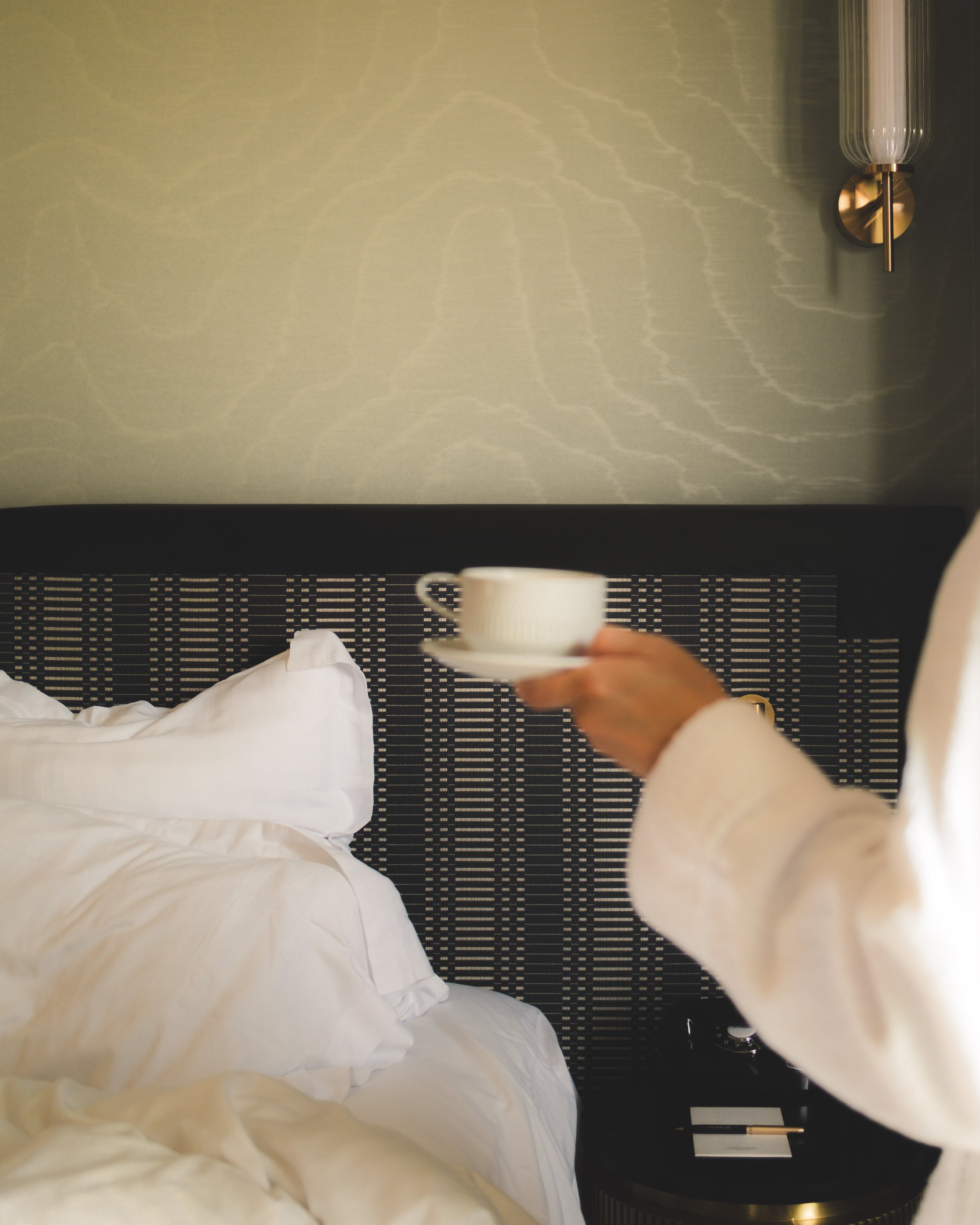 A hotel room with a bed covered in white linens, a tray with a white cup and saucer, a pen, keys, and a notepad on a black cabinet, a modern wall-mounted lamp, and a wall with a subtle textured pattern.