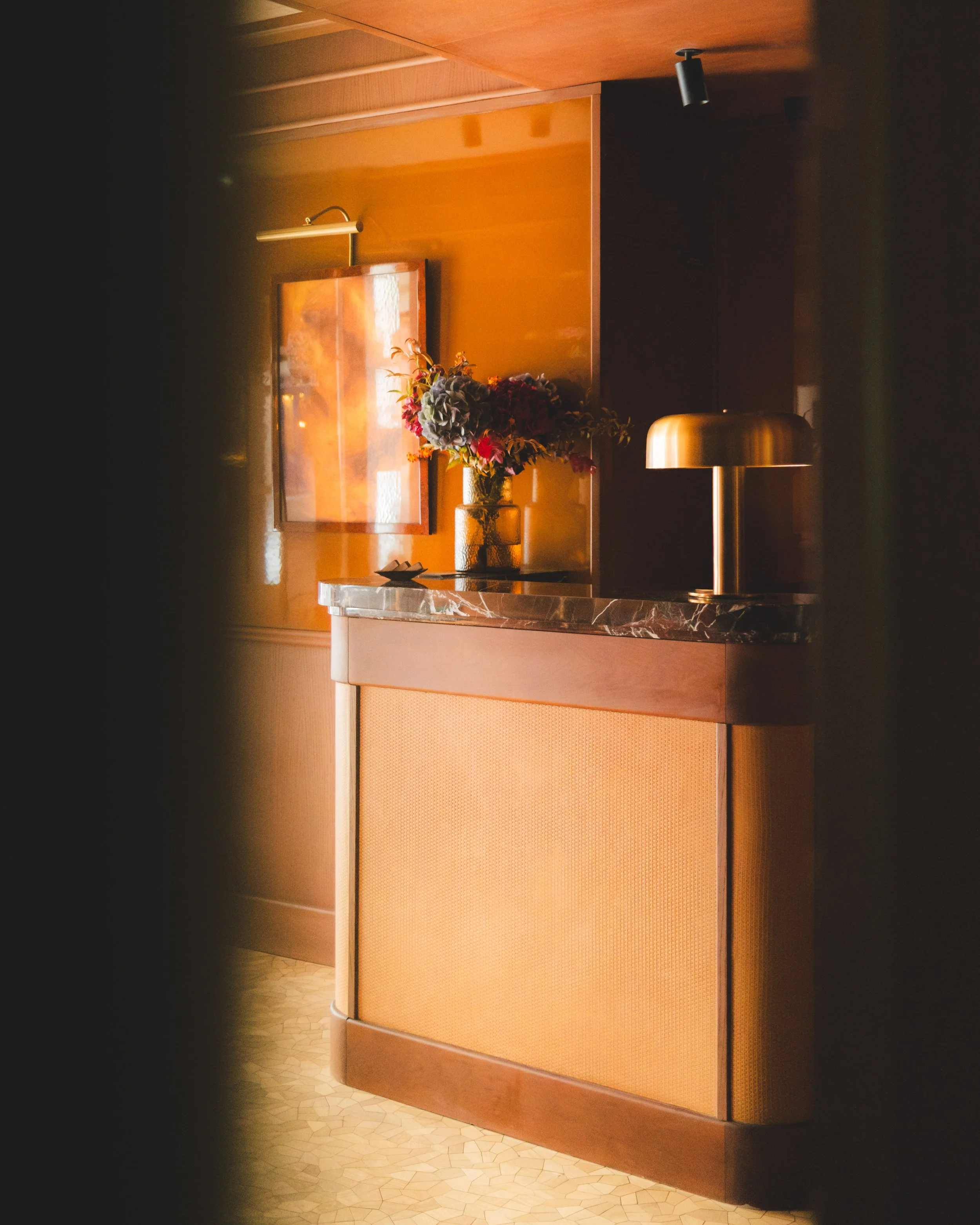 A corner of a room with a wooden wall, a mirror, a bouquet of flowers in a glass vase, a table with a marble top, and a modern lamp with a wide metallic shade.