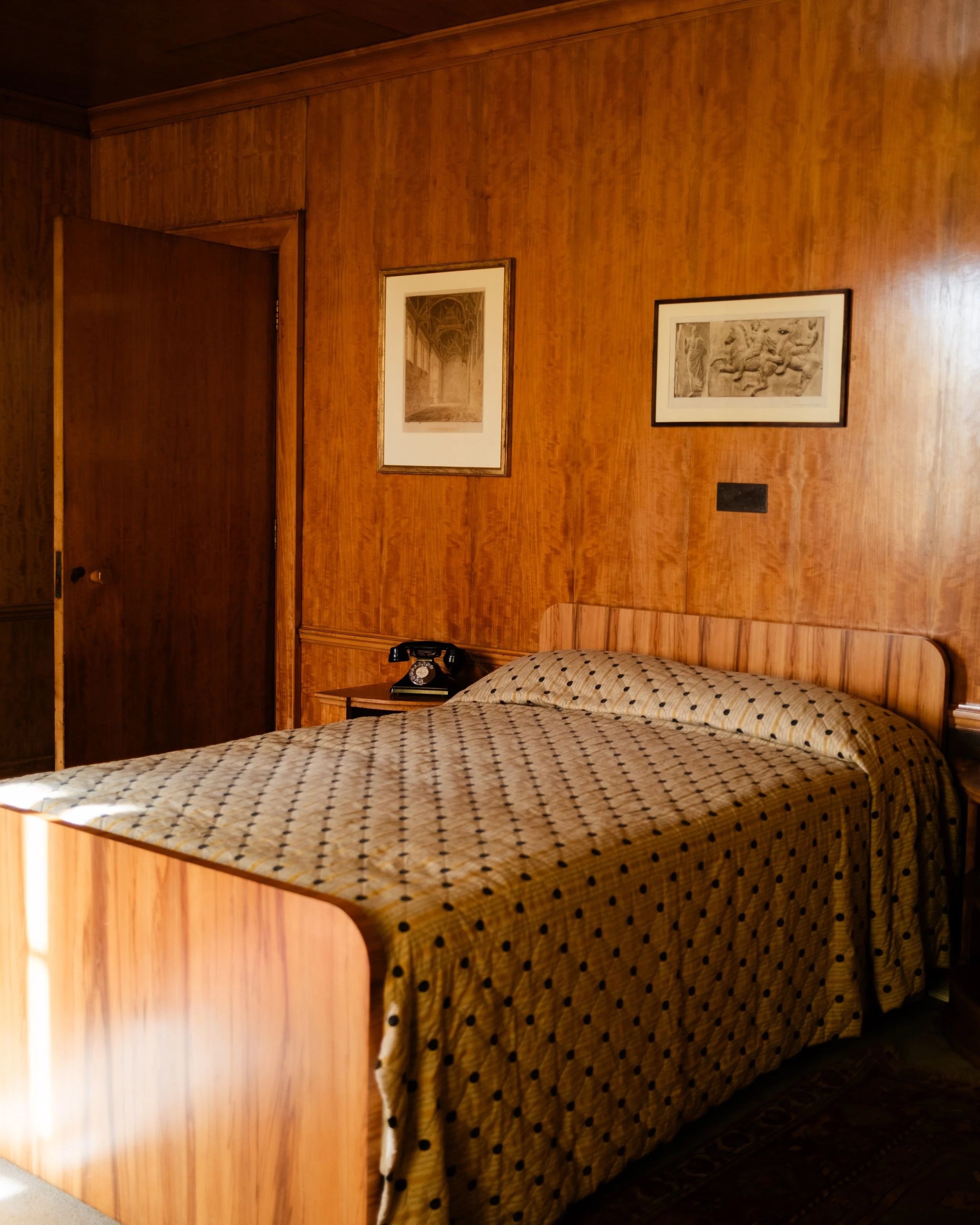 A vintage Bristish manor room with wood-paneled walls, a bed with polka dot bedding, a wooden headboard, a bedside table with a black rotary phone, and framed artwork on the walls. Lionel Chu - Hotel & Travel Photographer