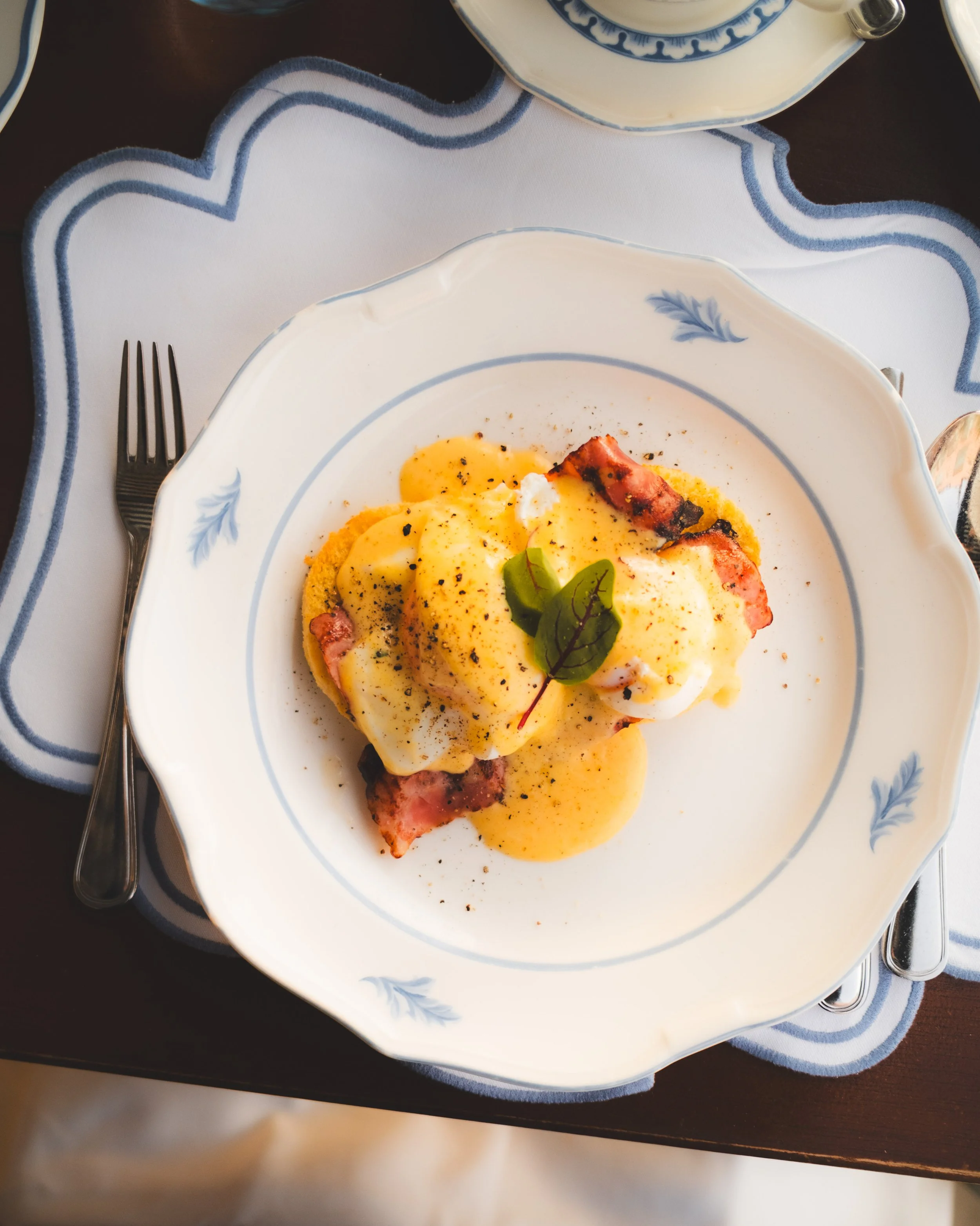 A plate of eggs Benedict with hollandaise sauce, slices of bacon, and a small green garnish on a decorative white and blue plate, set on a dark table with a white and blue-bordered placemat and a fork to the left.