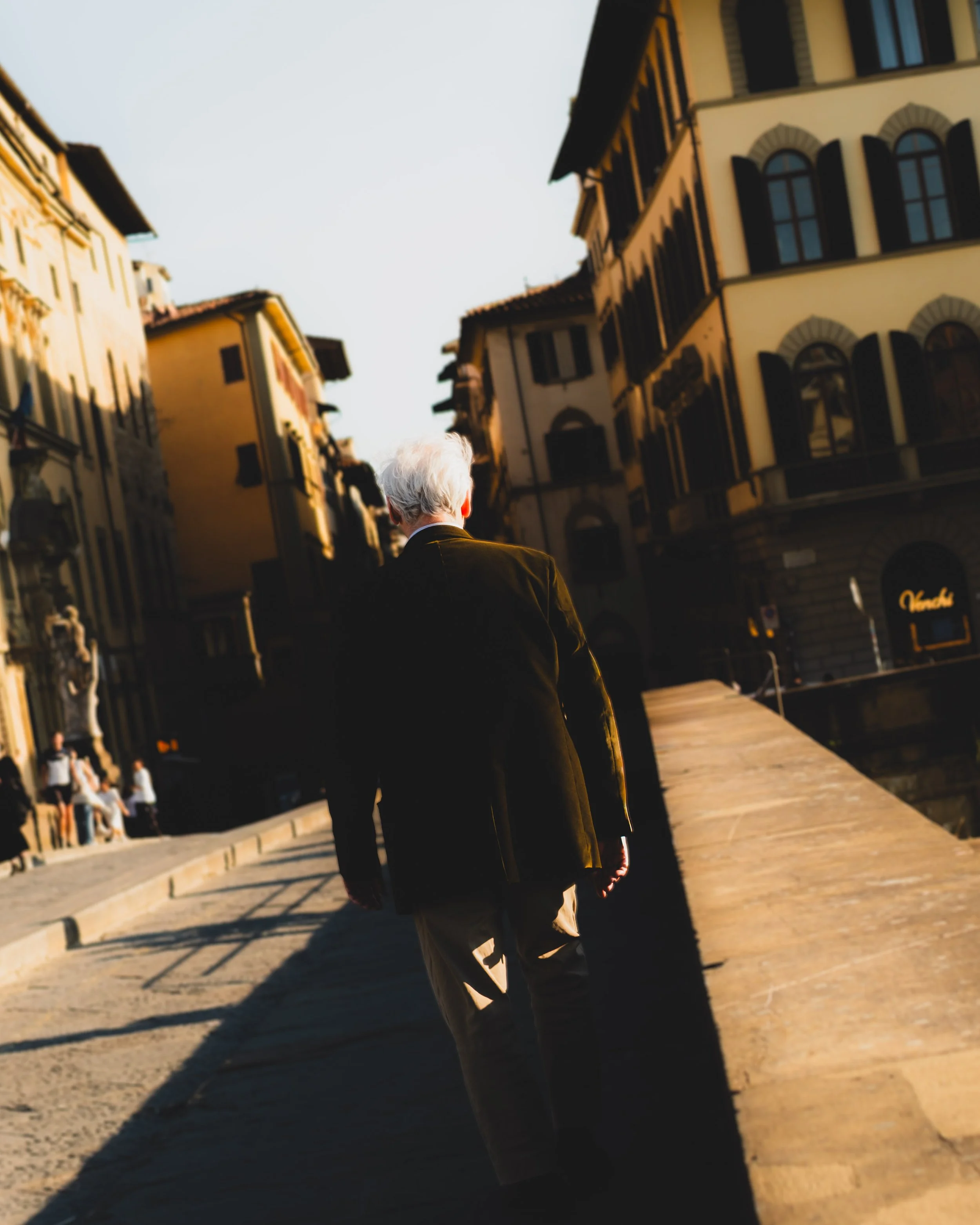 An elderly man with white hair, dressed in a dark jacket and light-colored pants, walking along a European city street during sunset, with old buildings and other pedestrians in the background. Lionel Chu - Hotel & Travel Photographer