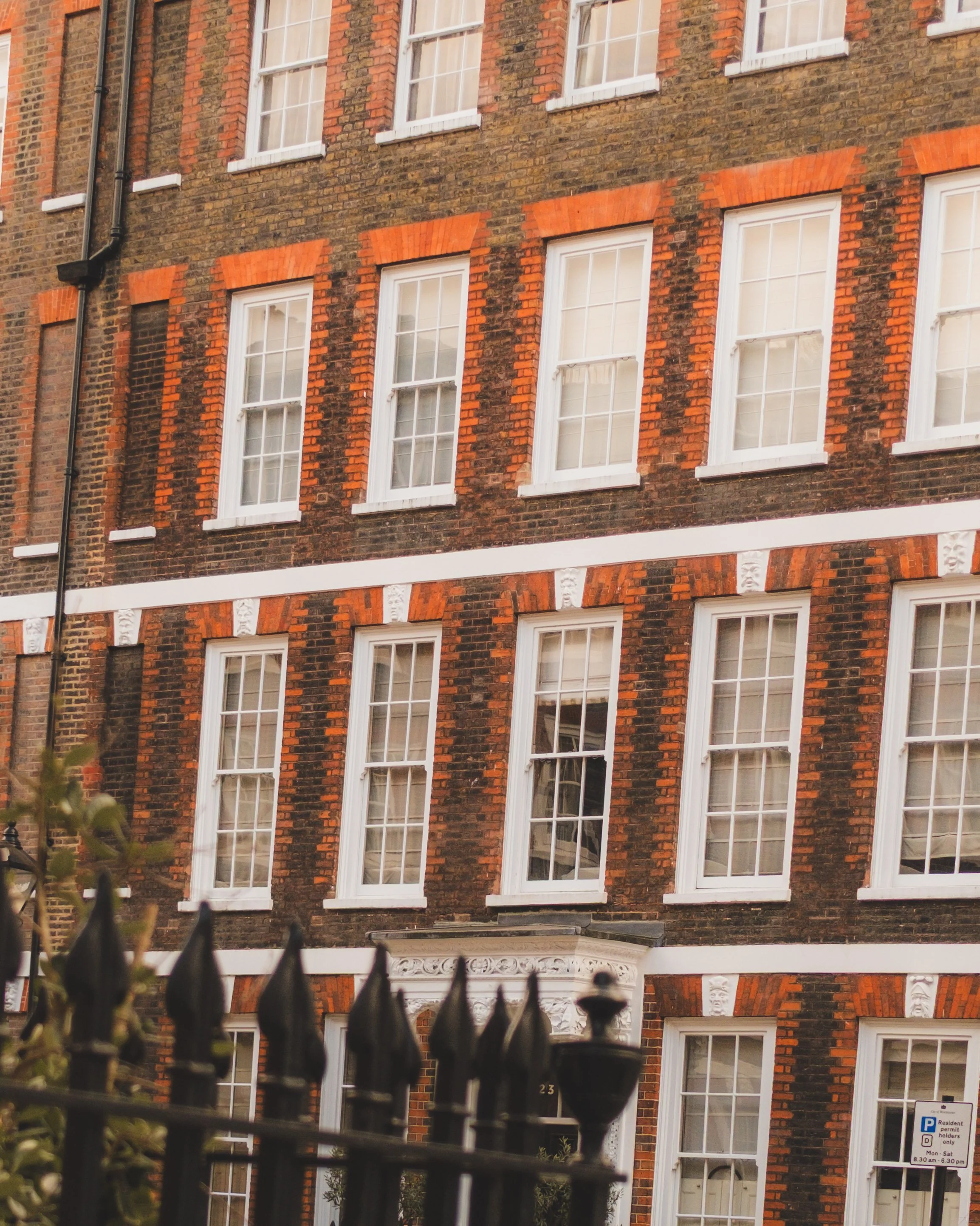A brick apartment building with multiple white-framed windows and decorative architectural details. A black metal fence is in the foreground.