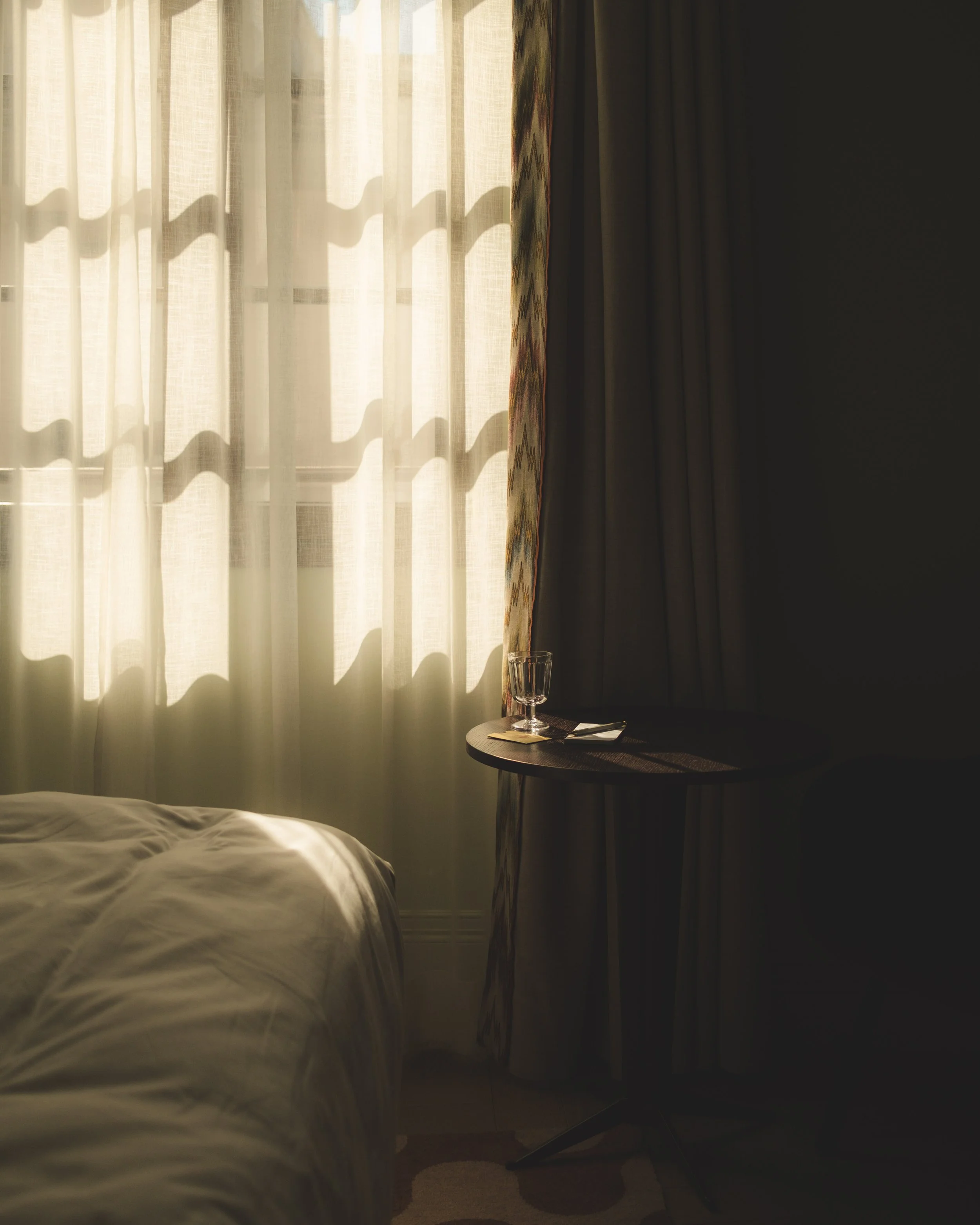 Sunlight filters through sheer curtains in a dimly lit room, with a small round table holding a glass of water and some utensils, partially visible, next to a bed.