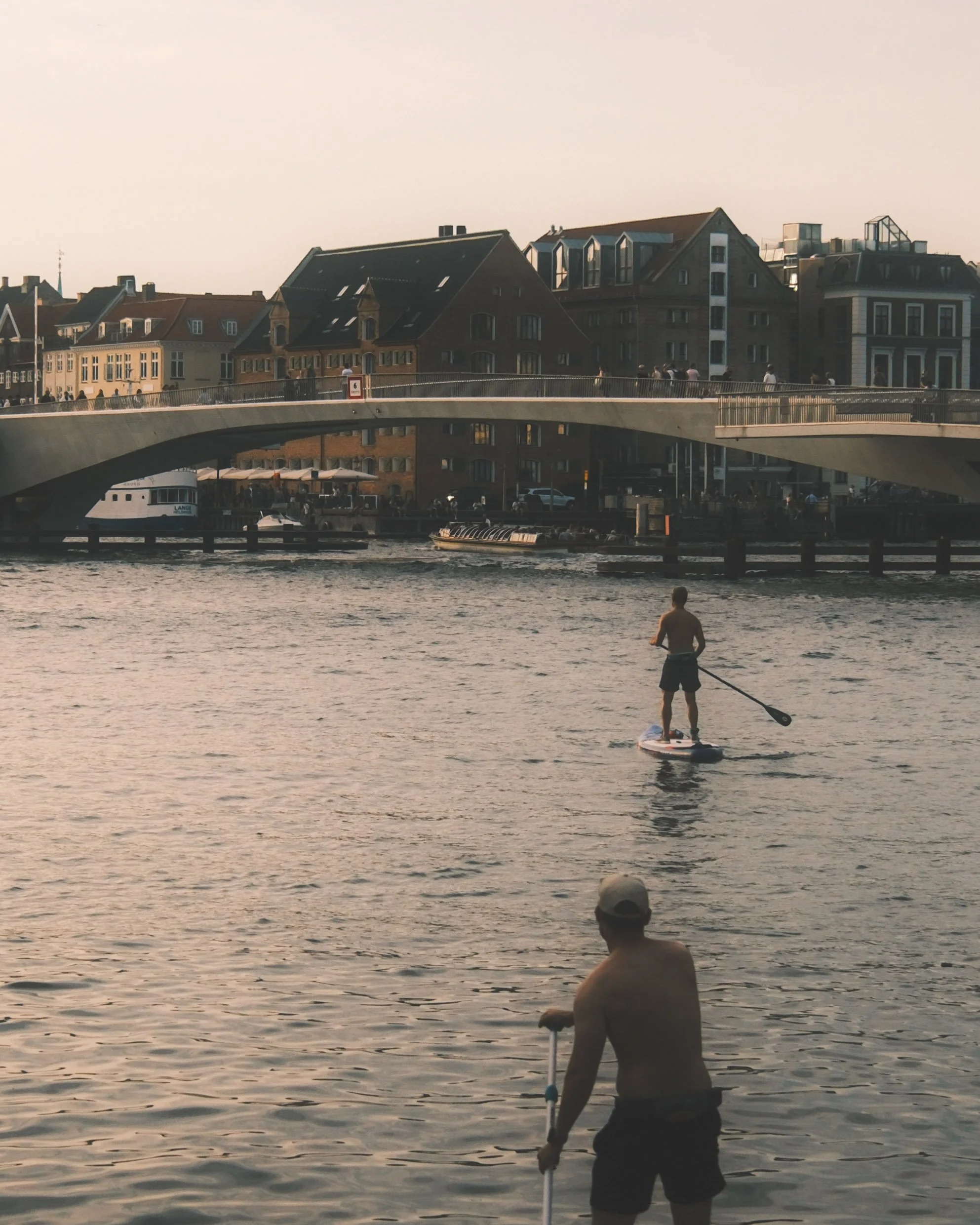 People enjoying a waterfront scene with a bridge, boats, and buildings in the background during the evening.