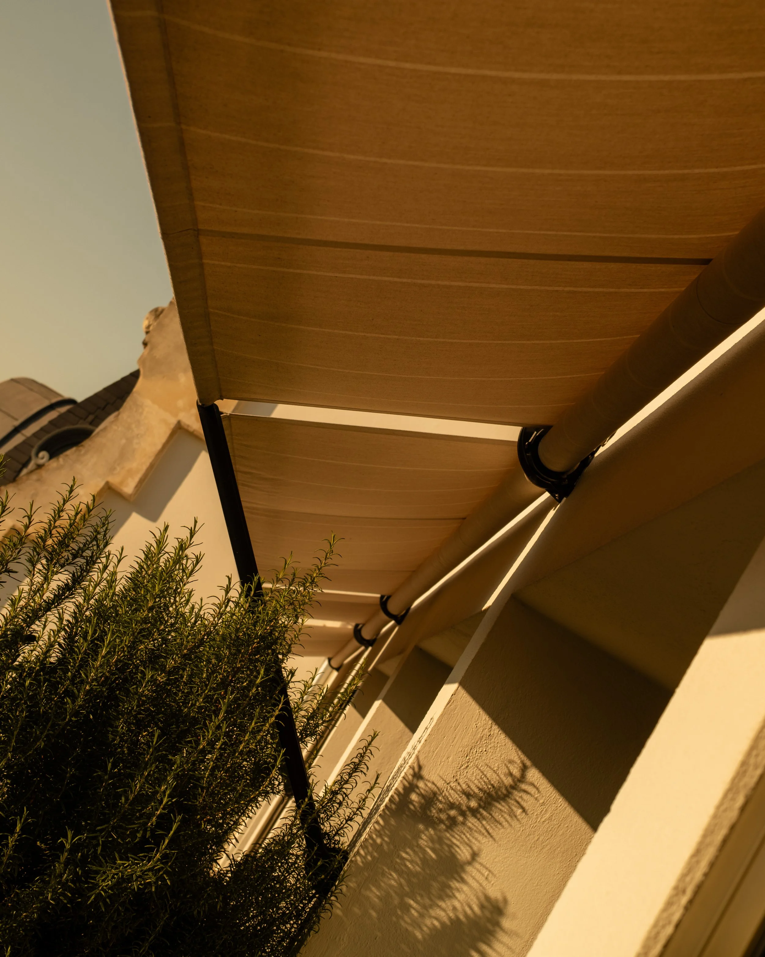 A photo taken from below showing a beige outdoor retractable awning, beige gutters, a beige wall, and a shrub with shadows cast on the wall. Lionel Chu - Hotel & Travel Photographer