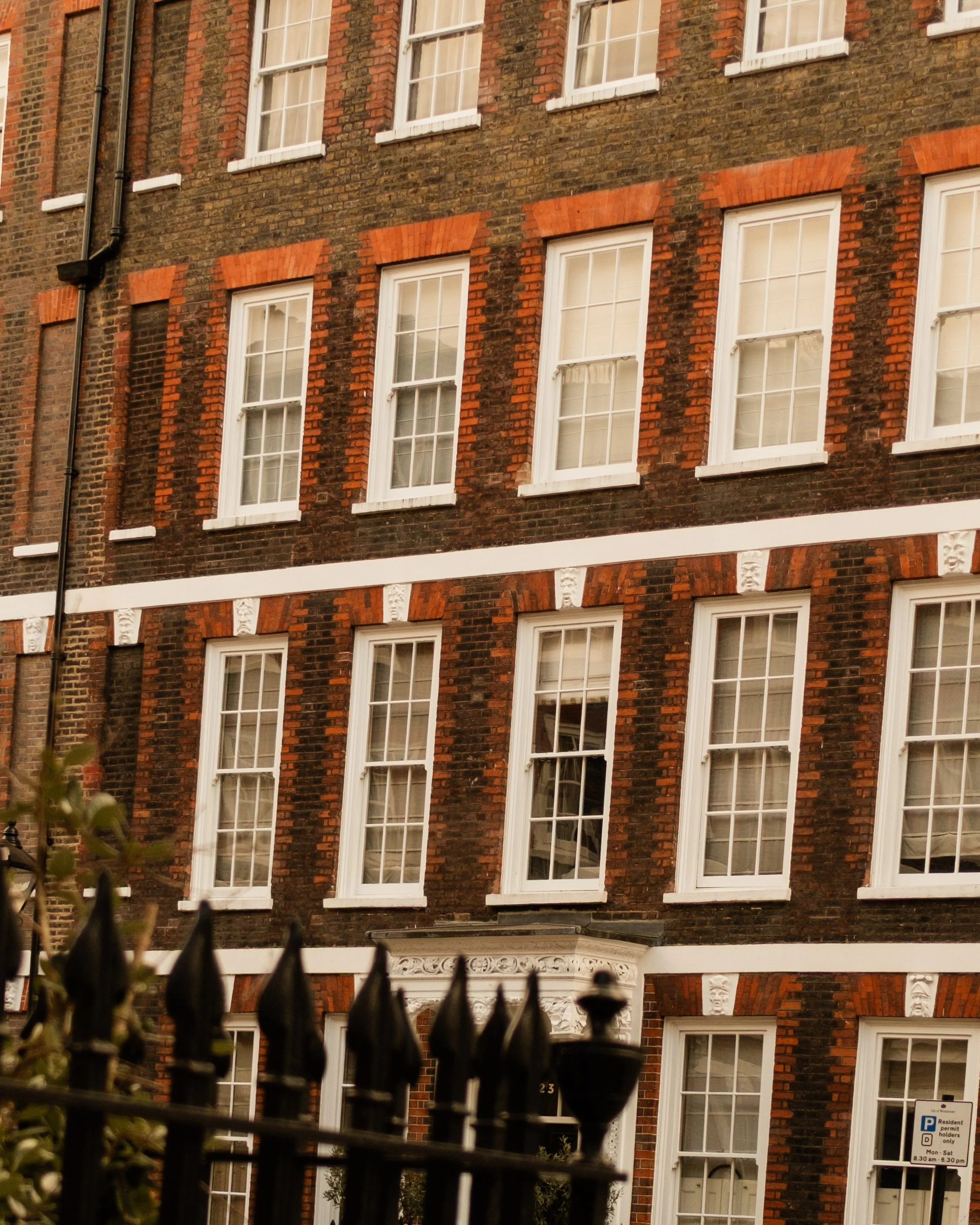 Three-story brick building with multiple white-framed sash windows, decorative white accents between floors, and a black iron fence at the front. The building has red brick arches above the windows and a black door with an ornate white decorative ped