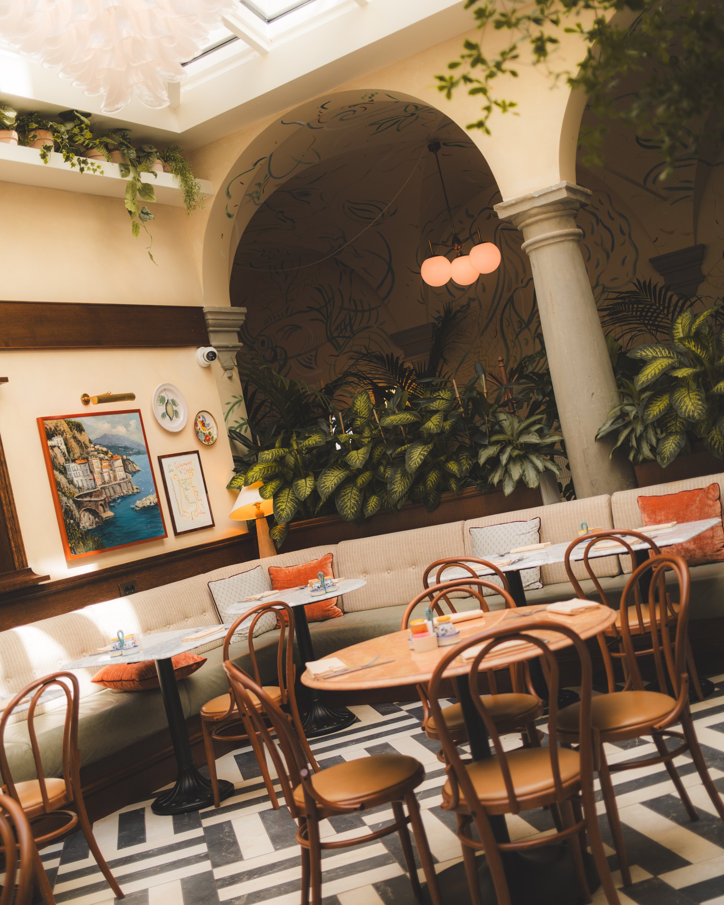 Interior of a cozy restaurant or cafe with a black and white checkered floor, tan upholstered bench seating with decorative pillows, round wooden tables, wooden chairs, leafy indoor plants, framed artwork on the wall, a decorative column, and a ceiling with a mural and a three-globe pendant light.