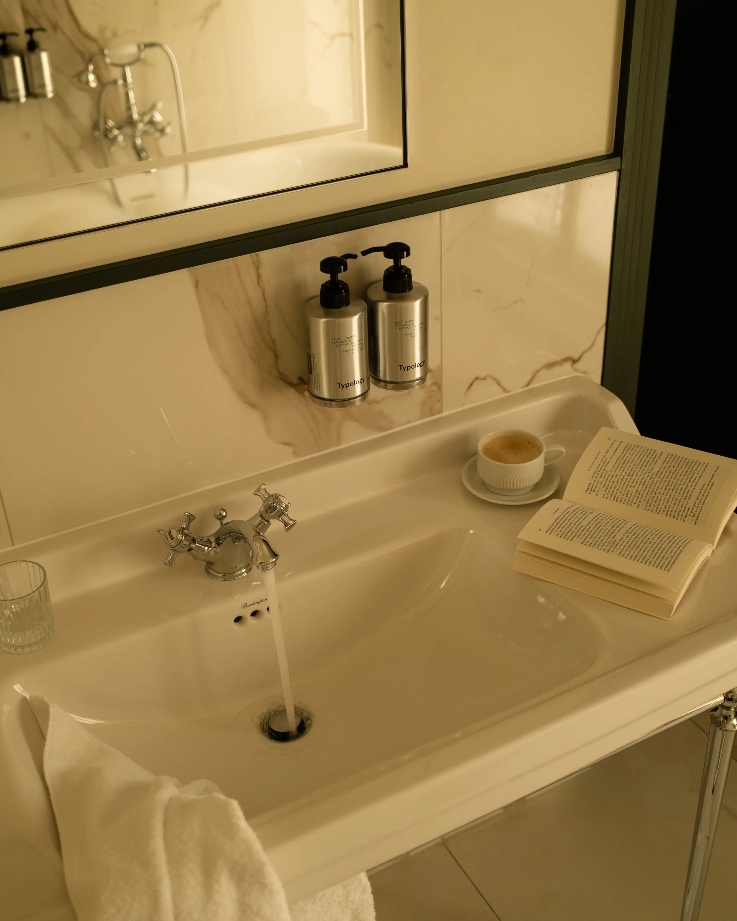 A bathroom sink with a cup of coffee, an open book, two soap dispensers labeled 'Typology,' and a small glass, with a mirror and wall-mounted fixtures in the background. Lionel Chu - Hotel & Travel Photographer