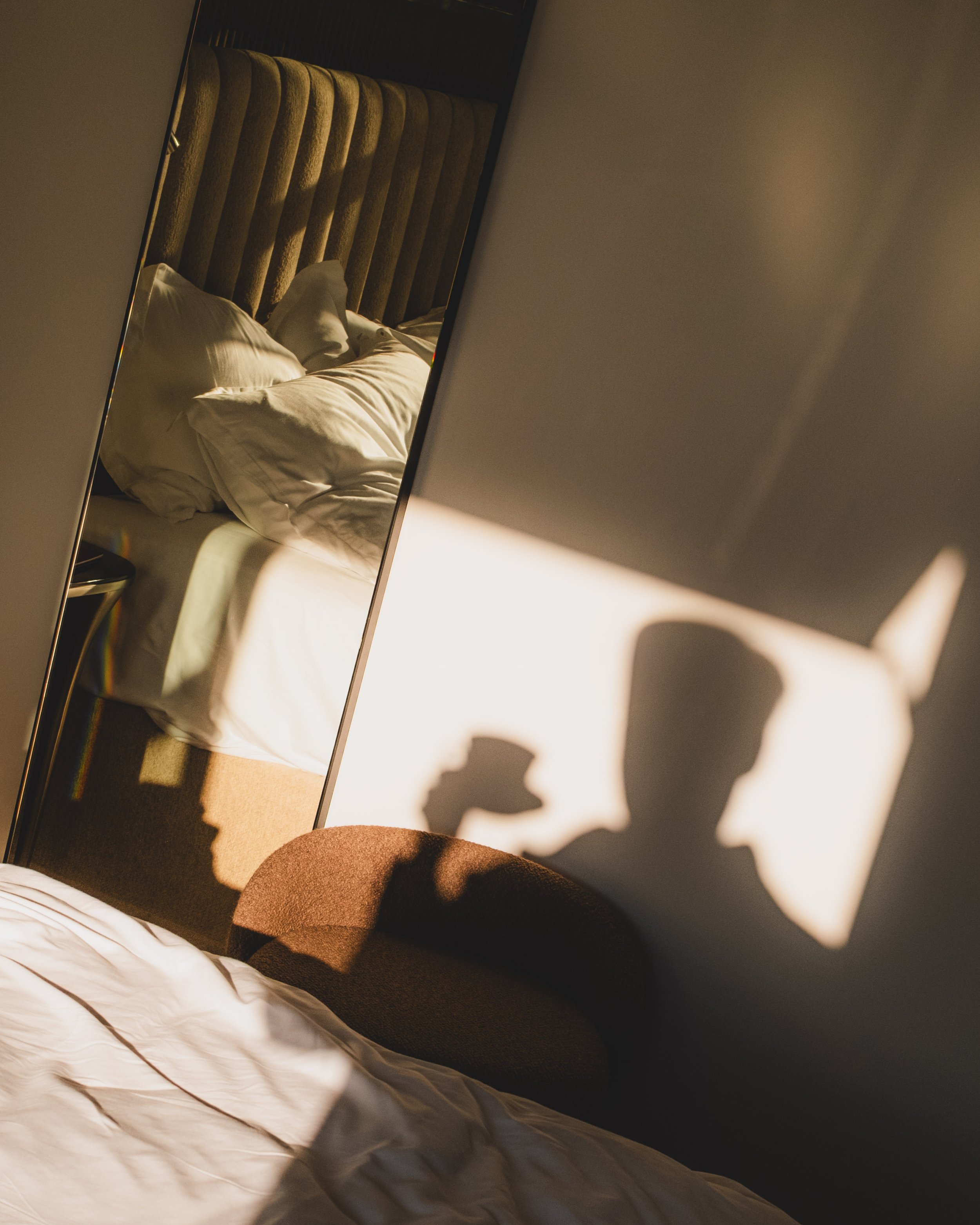 A mirror reflecting a bed with white sheets and pillows, a brown headboard, and a person wearing brown pants and white socks, capturing a cozy bedroom scene with dim lighting.