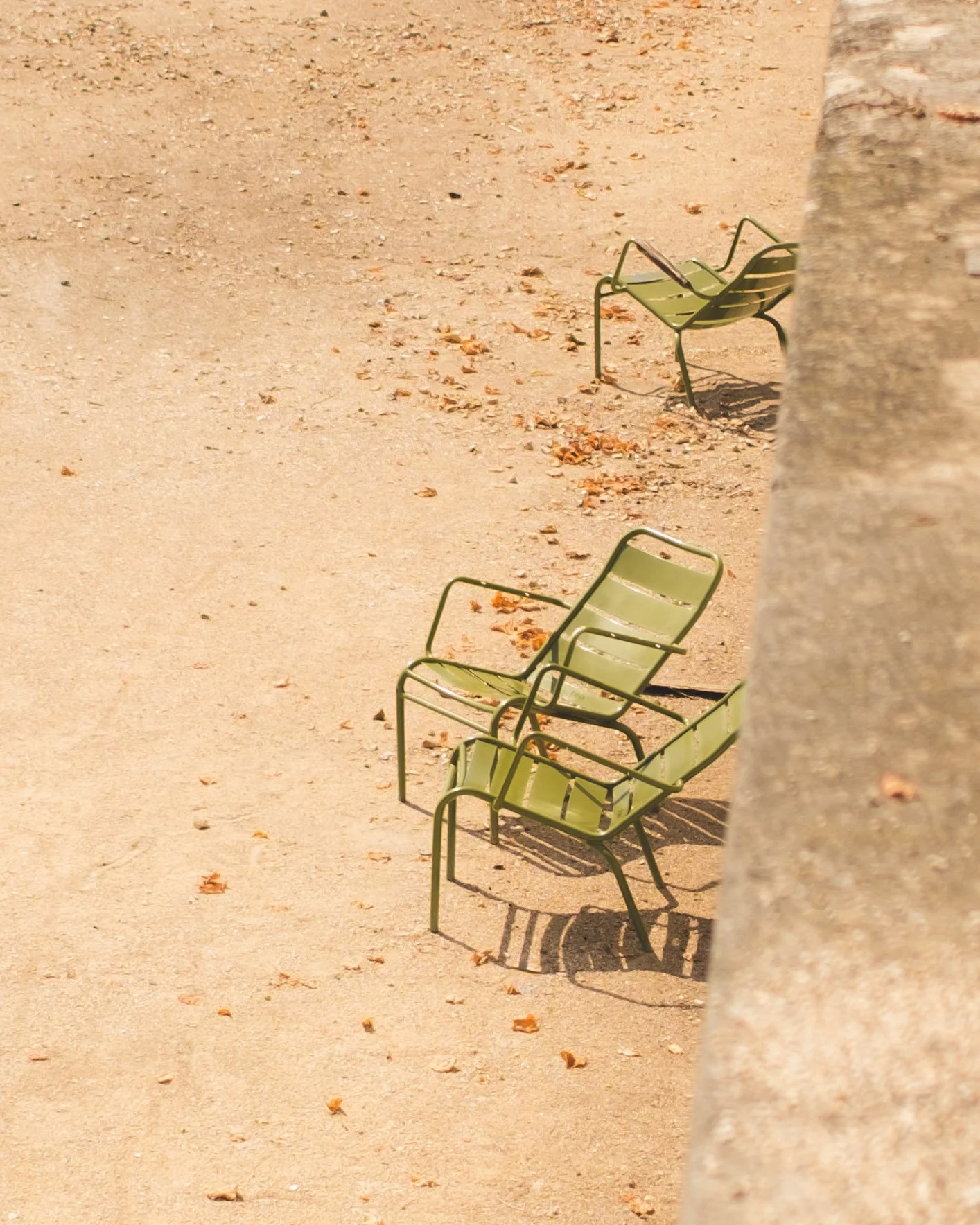 Three green metal chairs on a sandy ground with fallen leaves, partially obscured by a tree trunk in the foreground.