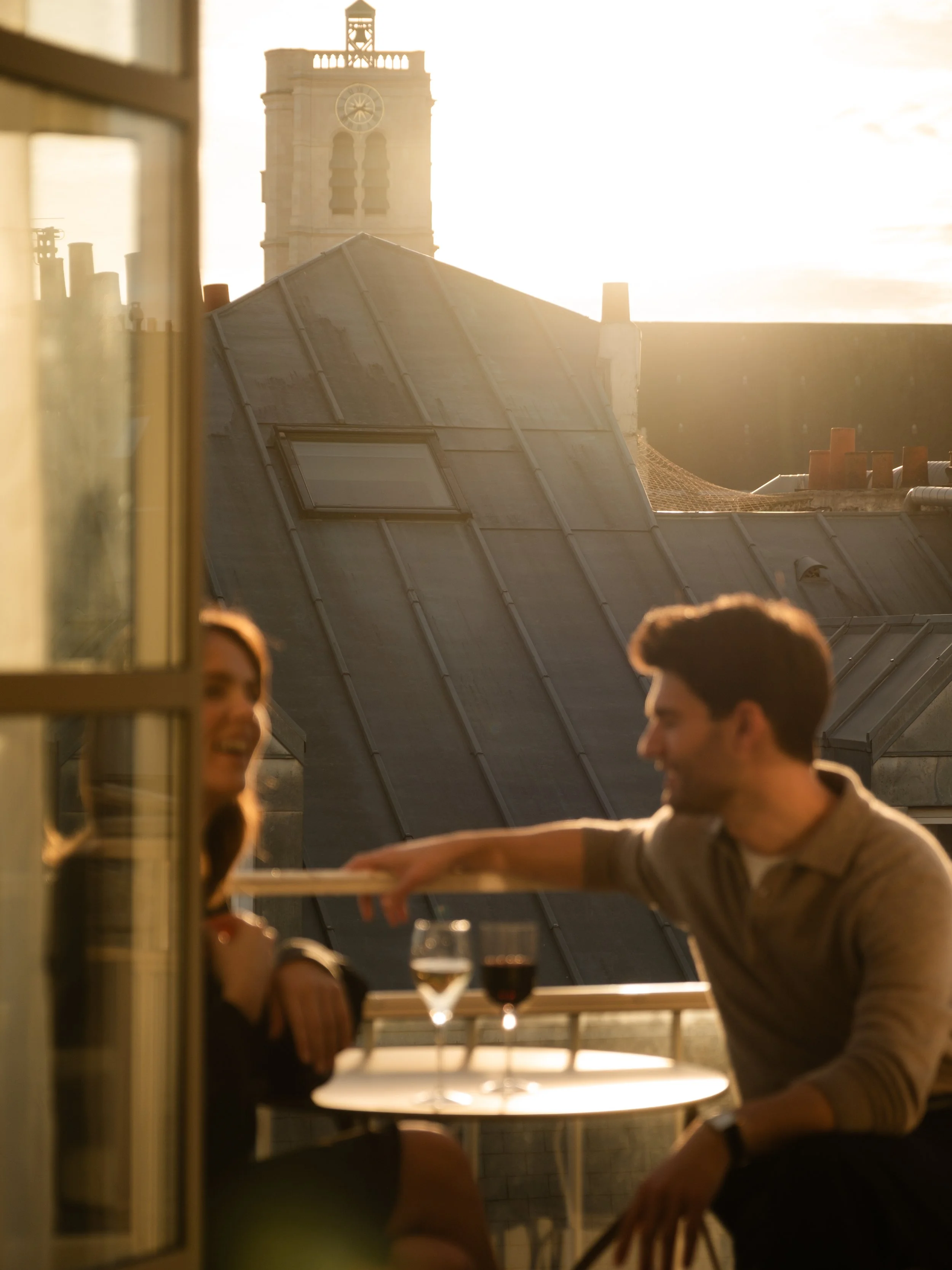 A man and woman sitting at a small table on a balcony, laughing and talking, with two glasses of wine in front of them, during sunset, with a church tower and rooftop structures in the background. Lionel Chu - Hotel & Travel Photography