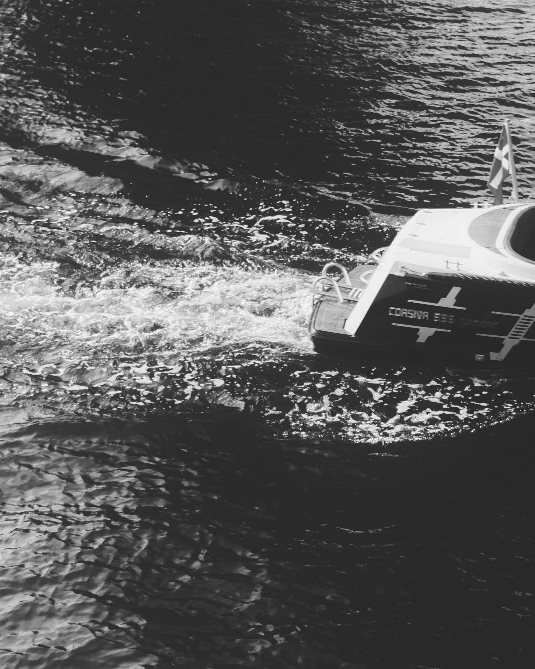 A small boat with a red and white flag on the stern, named Corsiva 595 Tender, moving through water, leaving a trail of waves behind. Lionel Chu - Hotel & Travel Photographer.