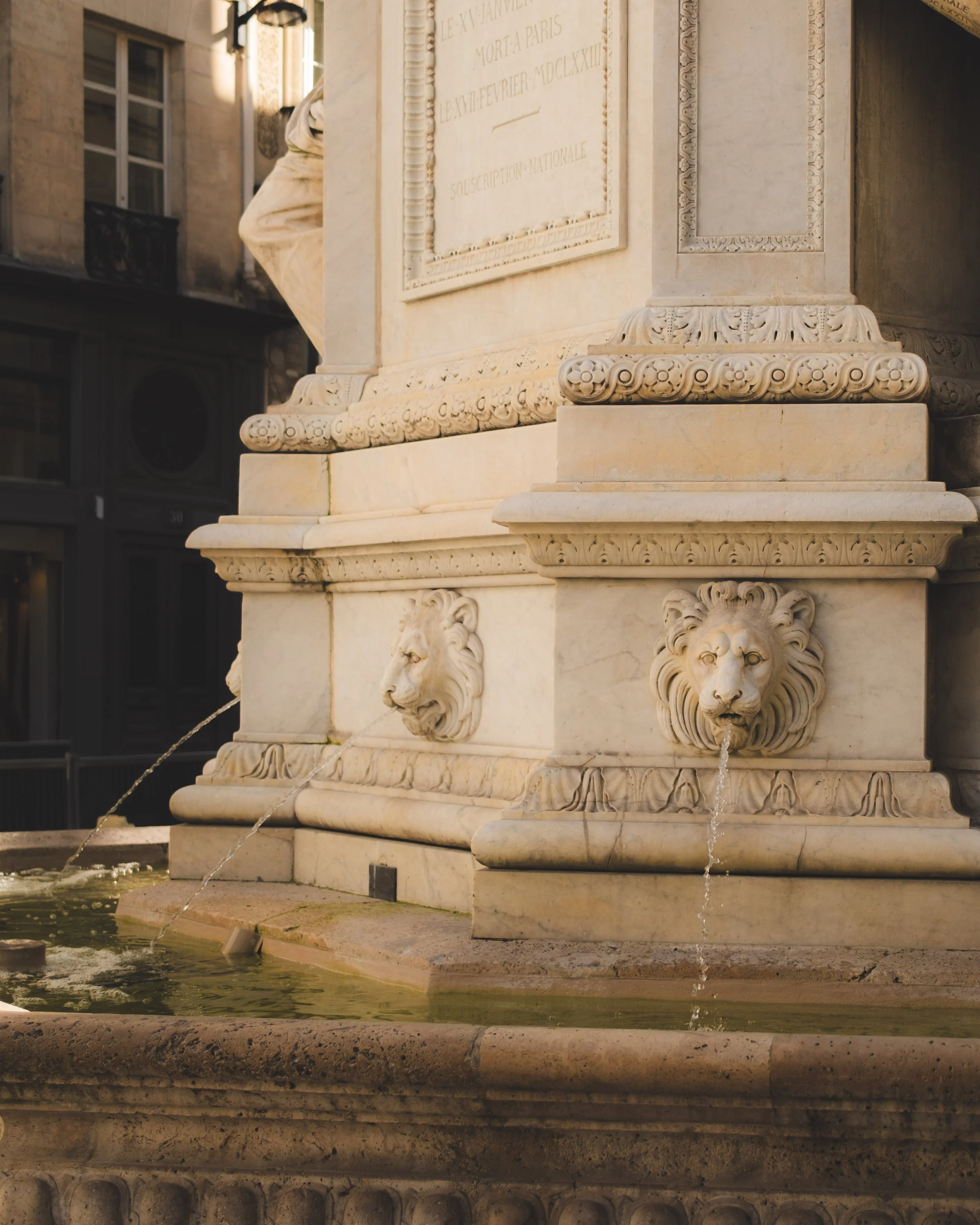 A marble fountain with three lion head sculptures, each with water flowing from their mouths, and an ornate square base decorated with carved floral and scroll motifs, located in an urban setting with buildings in the background.