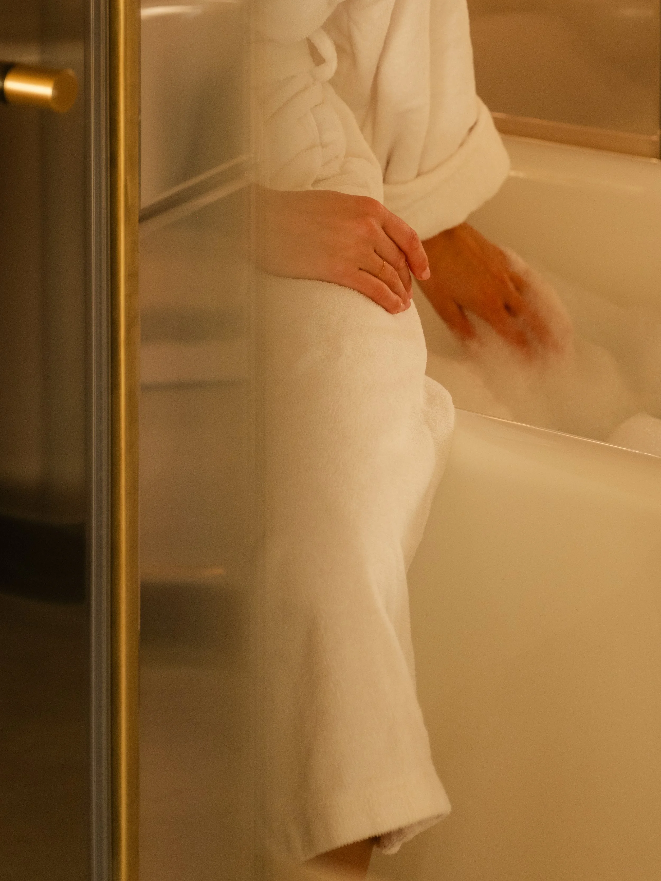 Person in a white bathrobe and towel sitting in a bathtub filled with bubbles, with hands resting on the lap, seen through a glass door. Lionel Chu - Hotel & Travel Photography