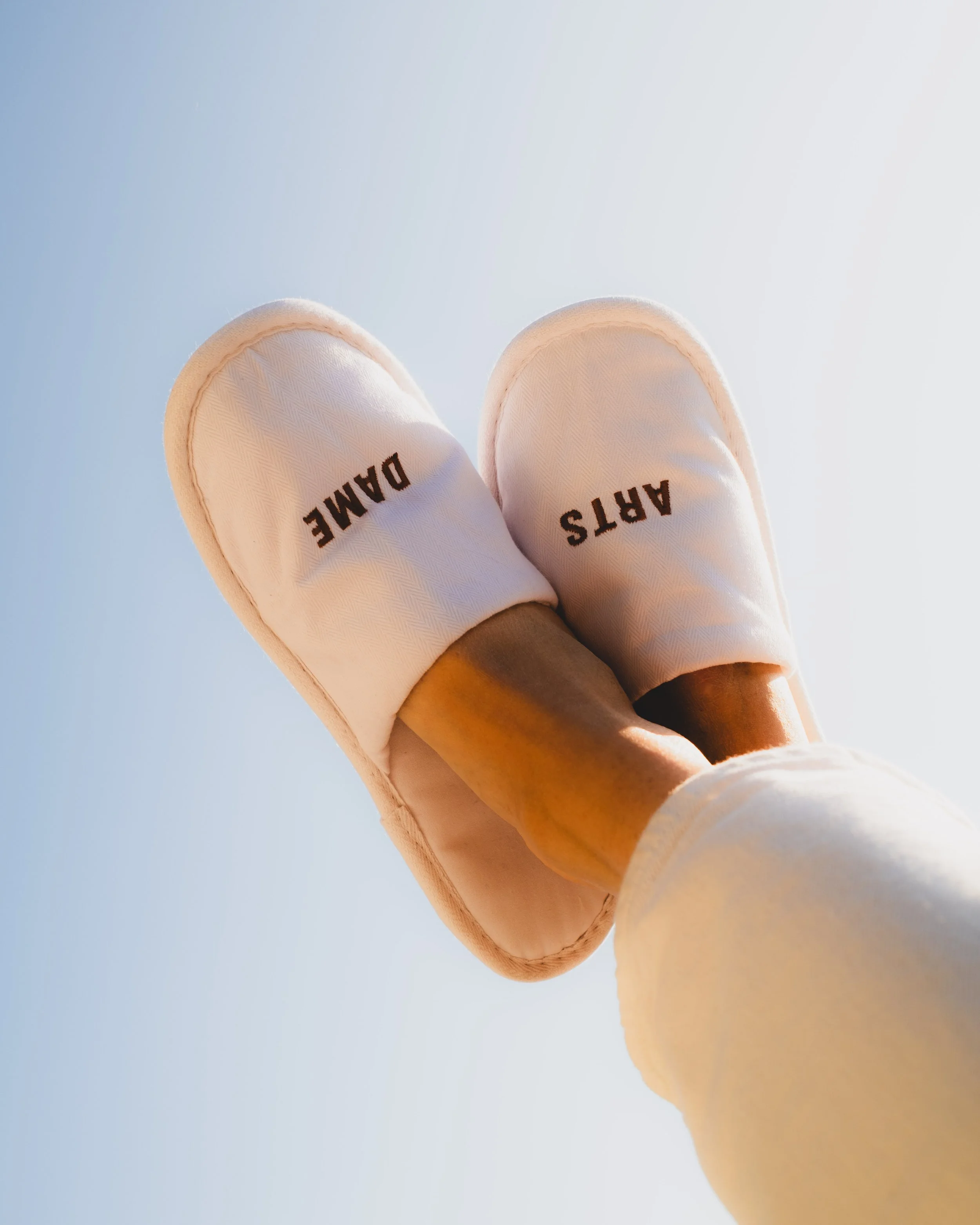 Close-up of a person wearing beige slippers with the word 'ARTS' on one slipper and 'DAME' on the other, against a clear blue sky.