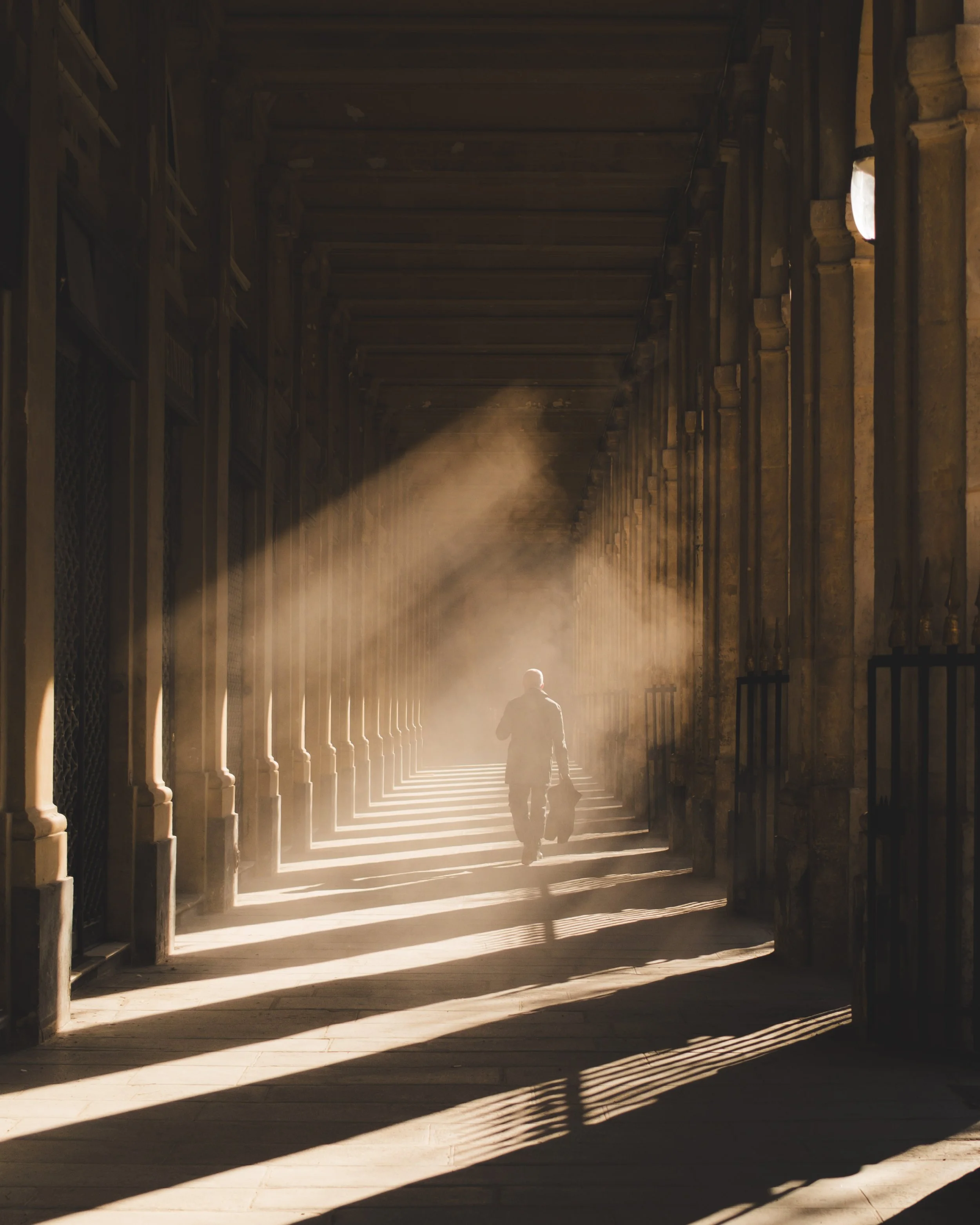 A person walking through a historic arched corridor illuminated by sunlight, carrying a bag.