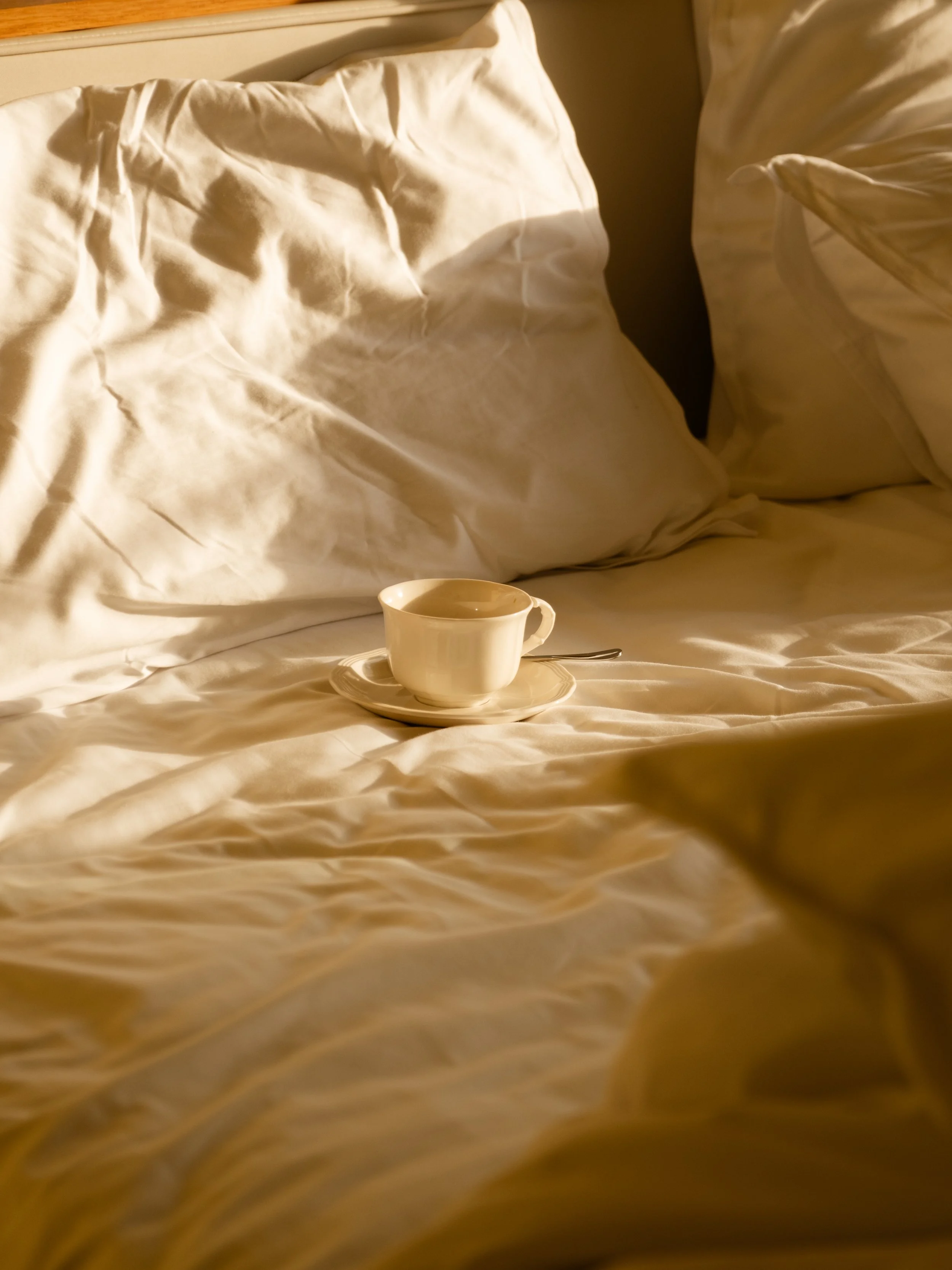 A white ceramic coffee cup with a saucer and spoon placed on a bed with white sheets and pillows.
