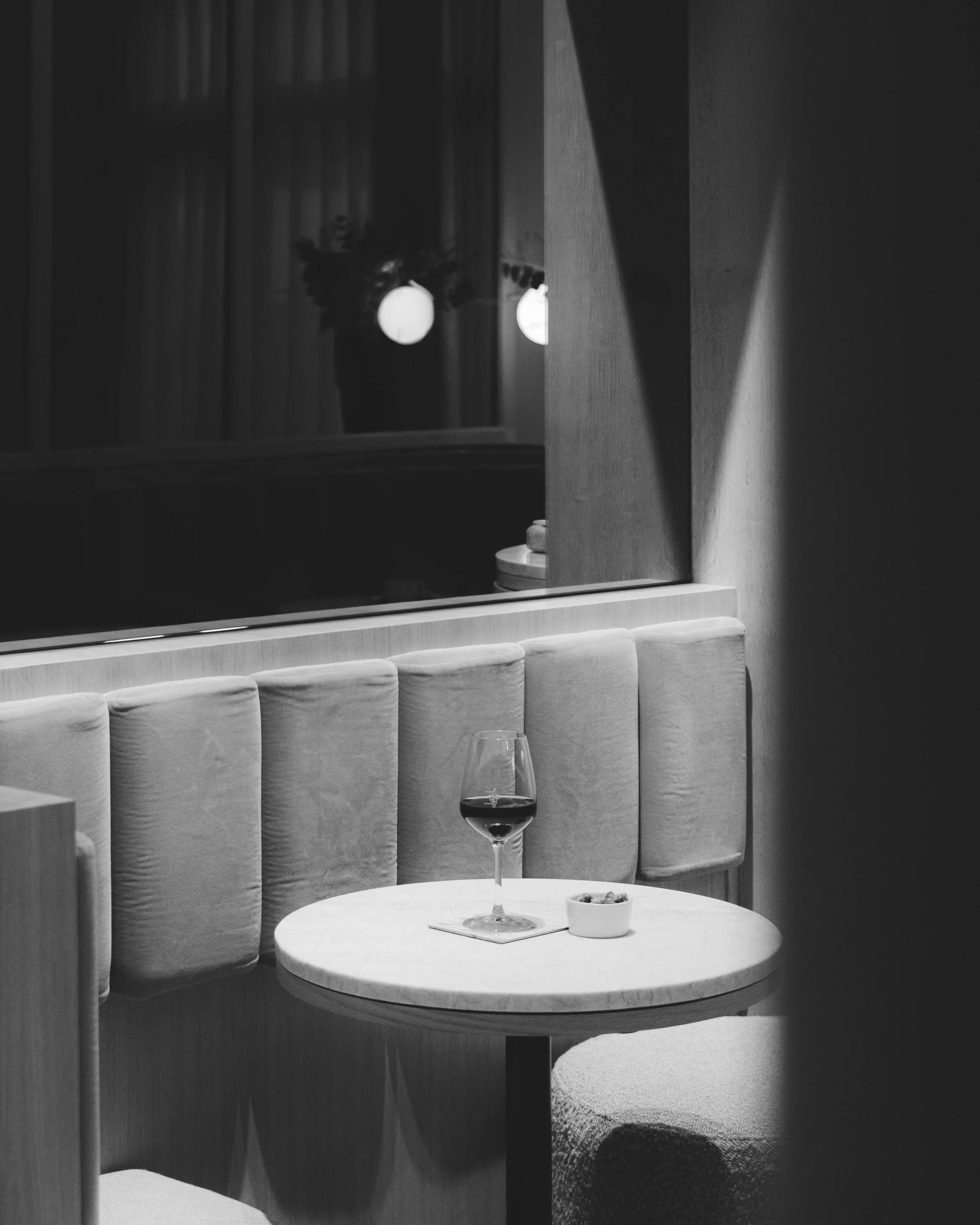 Black-and-white photograph of a dining scene with a round table, a glass of red wine, a small bowl of snacks, and upholstered chairs. In the background, a mirror reflects a window with curtains and a floral arrangement with spherical light fixtures.
