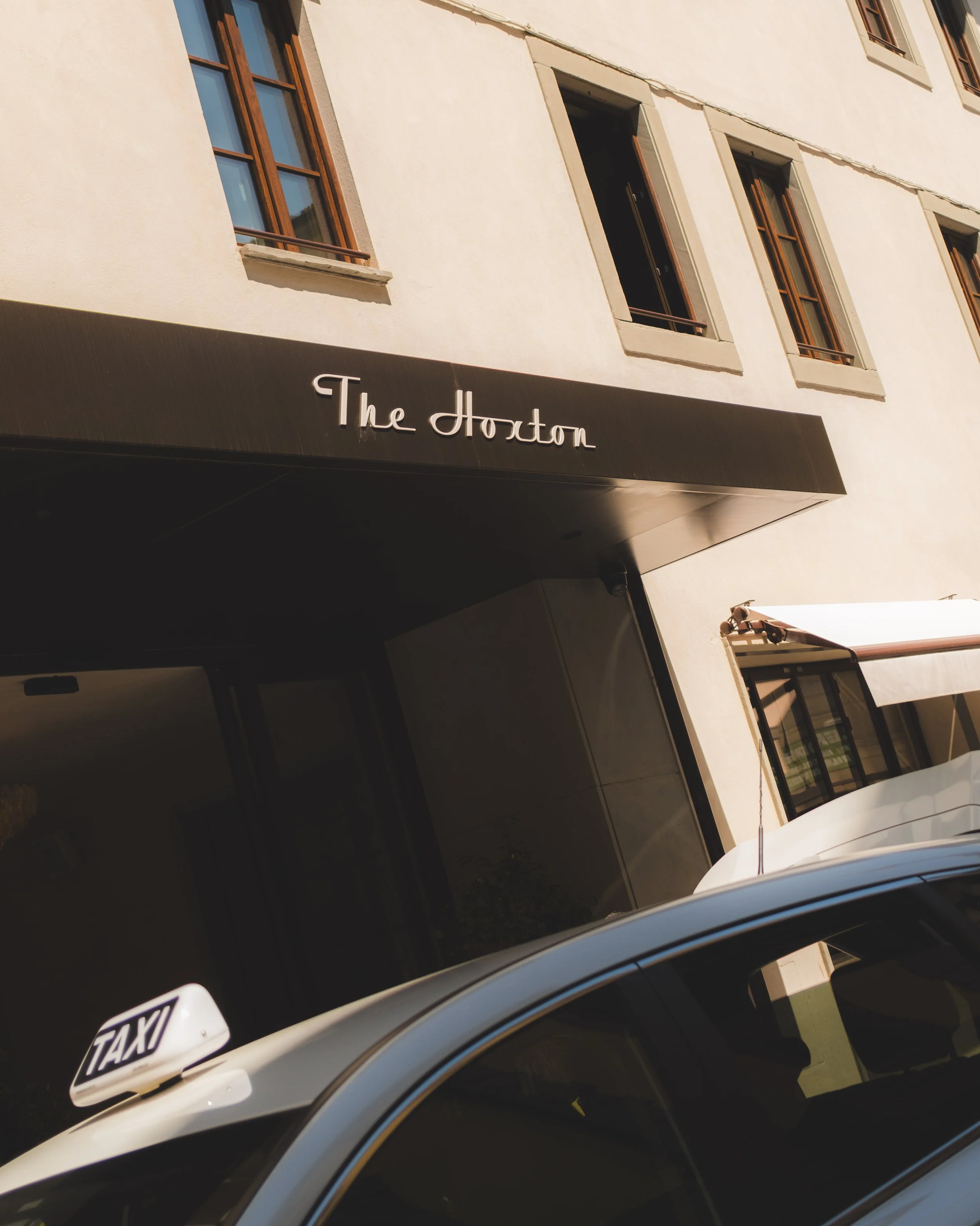 The image shows the side of a building with several windows and a black awning with the white text 'The Hoxton'. In the foreground, there is a white taxi with a visible part of the taxi sign on the roof, and a partially visible license plate.