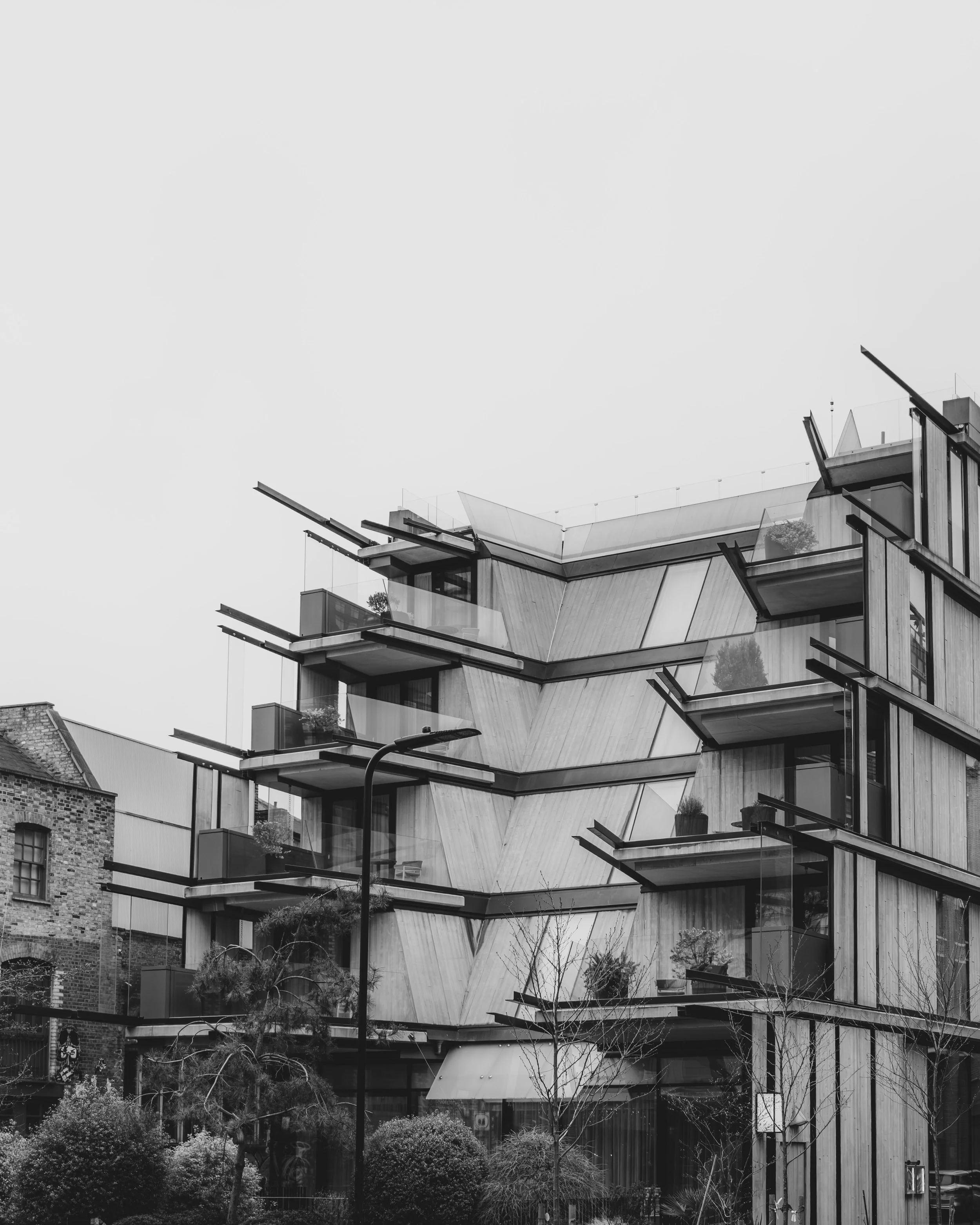 Modern multi-story building with angular glass balconies and wooden paneling, with trees and bushes in the foreground.