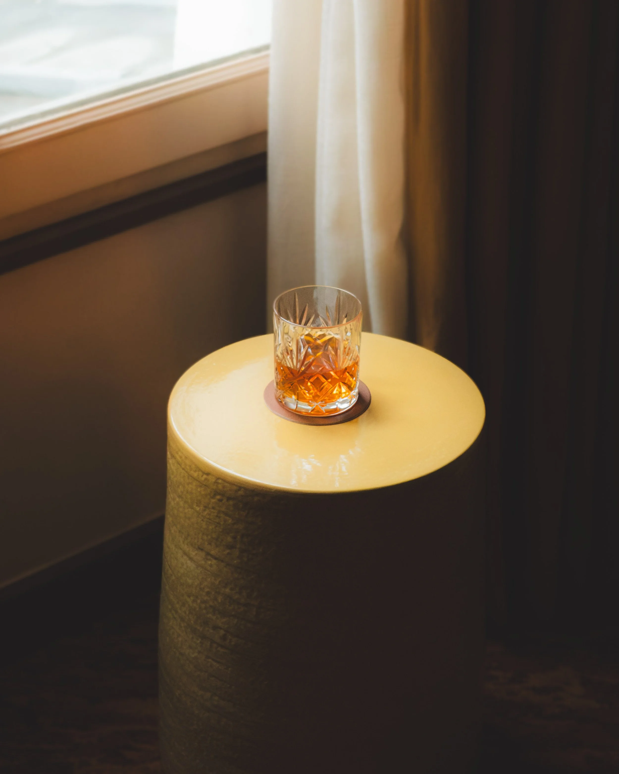A glass of amber-colored drink on a small round table near a window with curtains.
