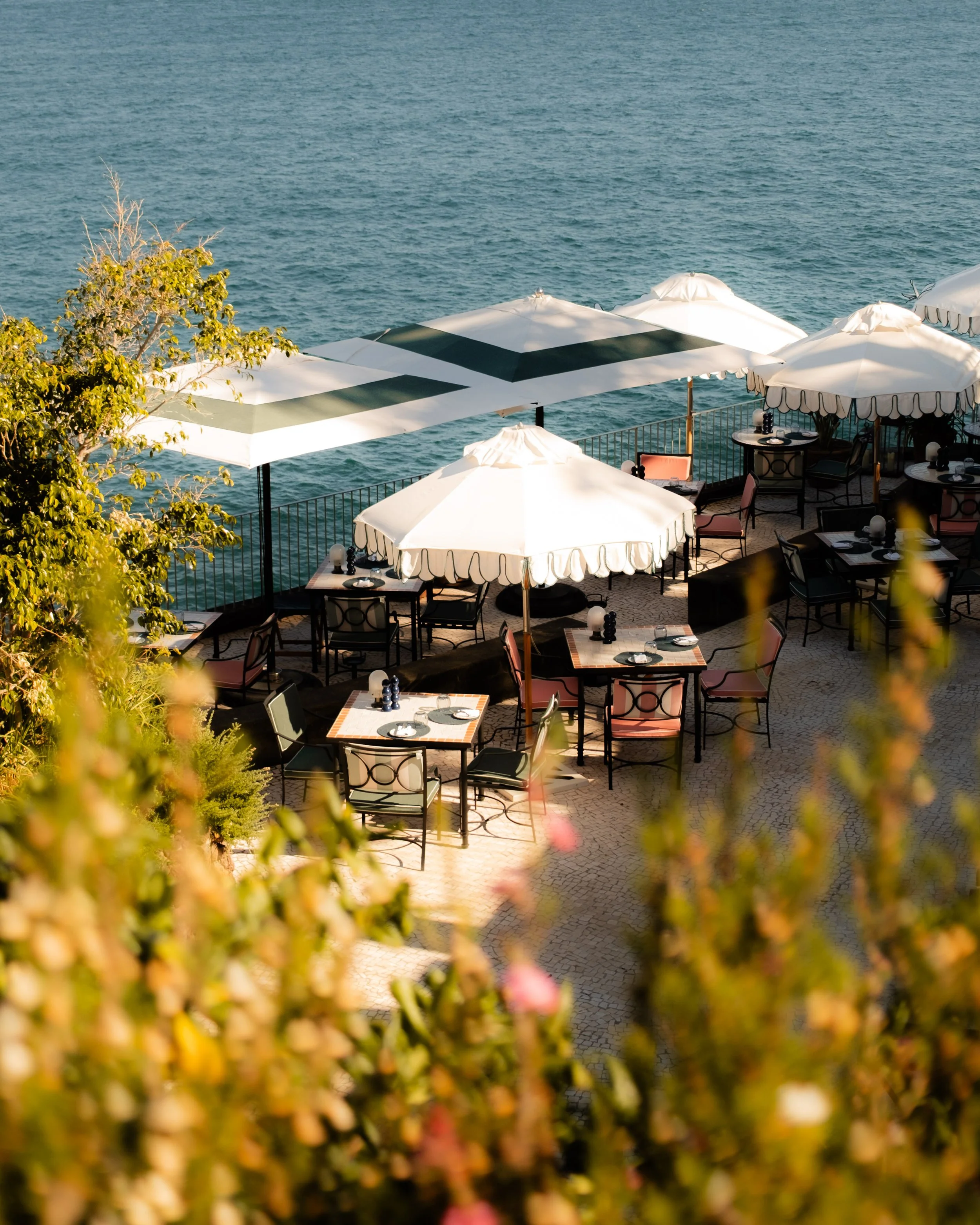 Outdoor seating area with tables, chairs, and large white umbrellas overlooking a body of water, with some greenery in the foreground.