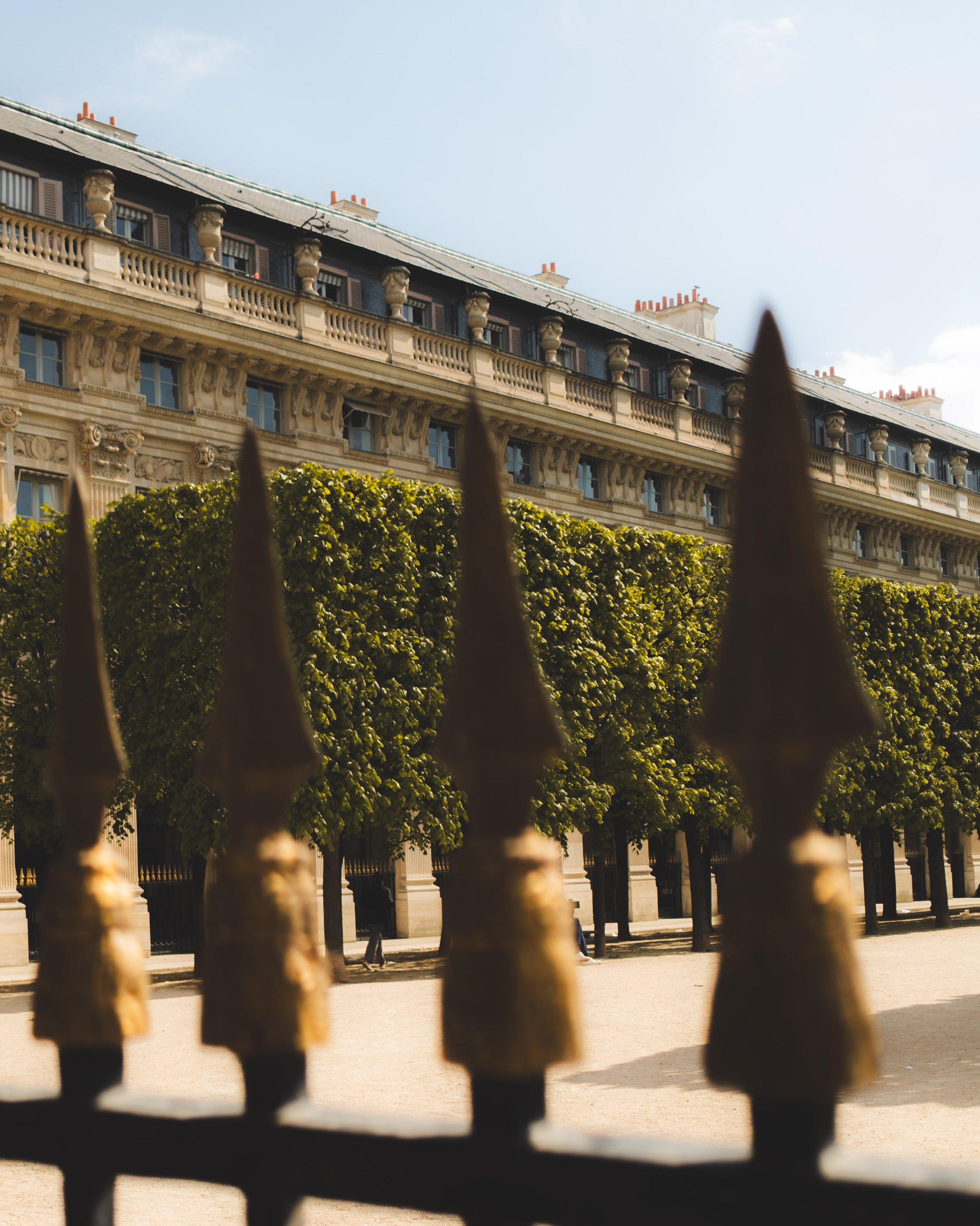 View of the Palais Royal Garden in Paris with ornate architecture, seen through an iron fence with pointed tips in the foreground, under a clear sky.