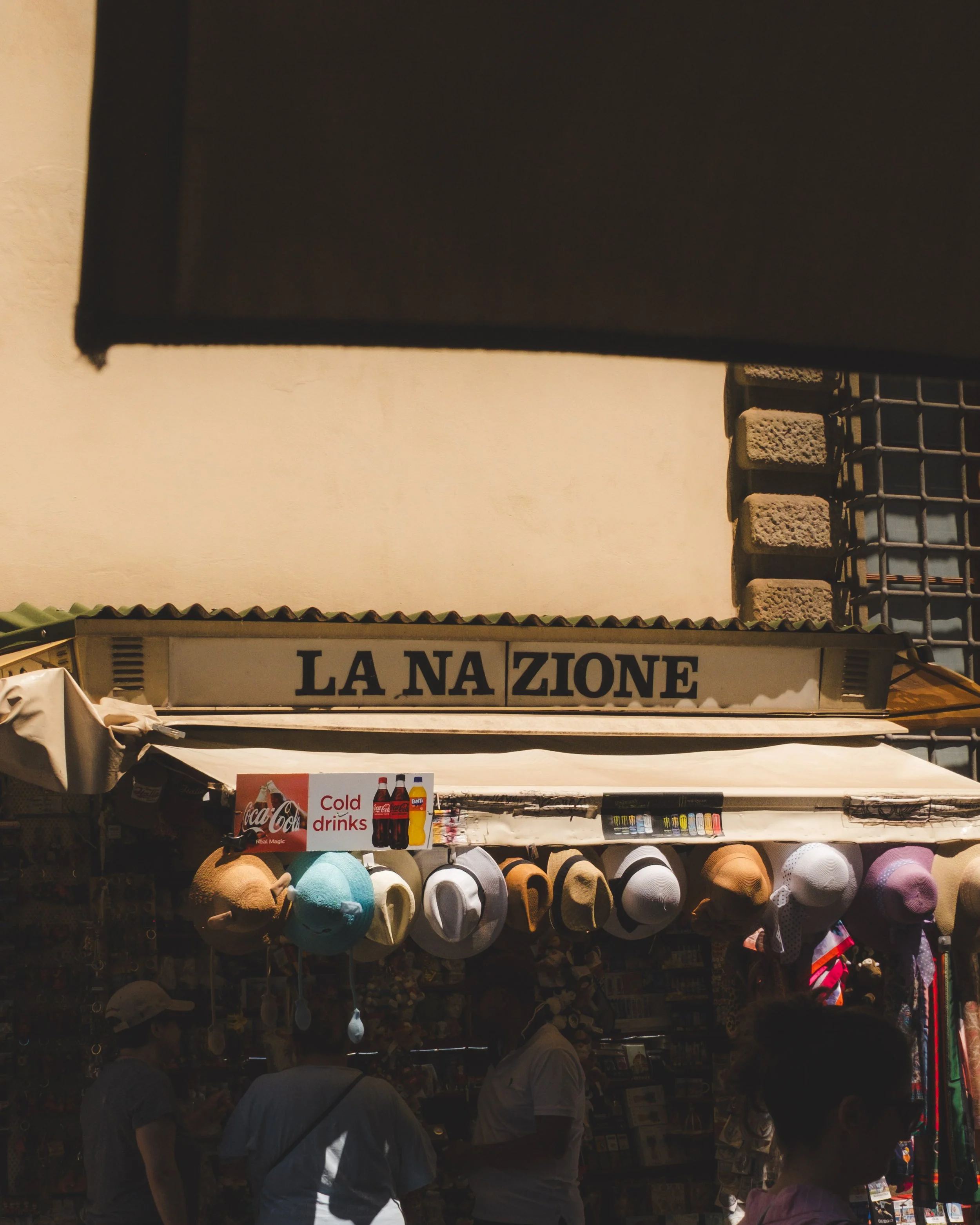 A street stall with hats and signs, including one for cold drinks, located under a sign that reads 'LA NAZIONE' in an outdoor market scene. Lionel Chu - Hotel & Travel Photographer