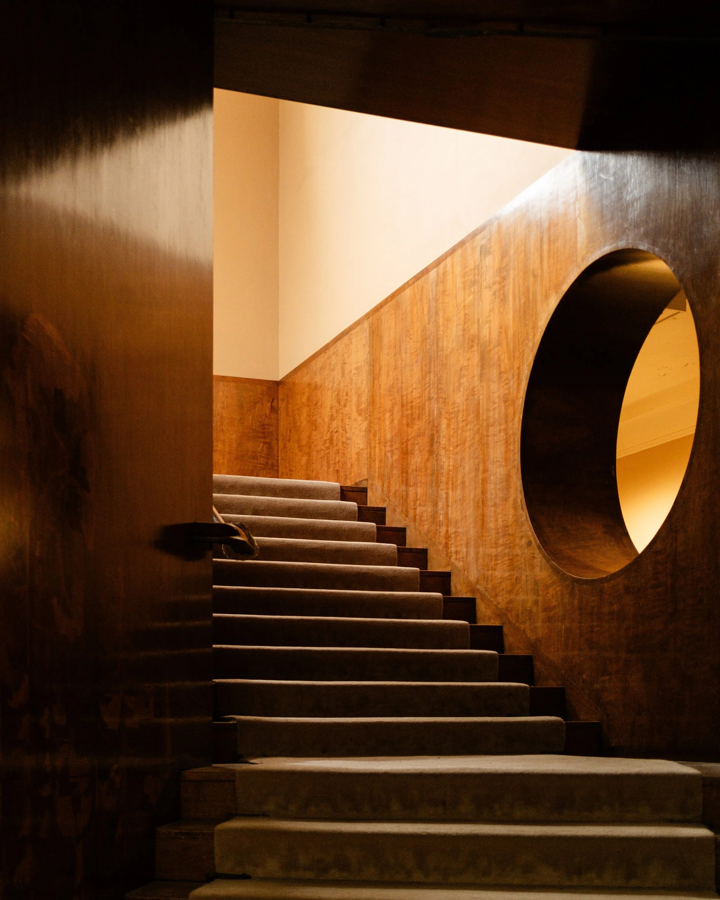 A staircase with wooden walls, carpeted steps, and a large circular cutout in the wall, viewed from the bottom of the stairs. Lionel Chu - Hotel & Travel Photographer