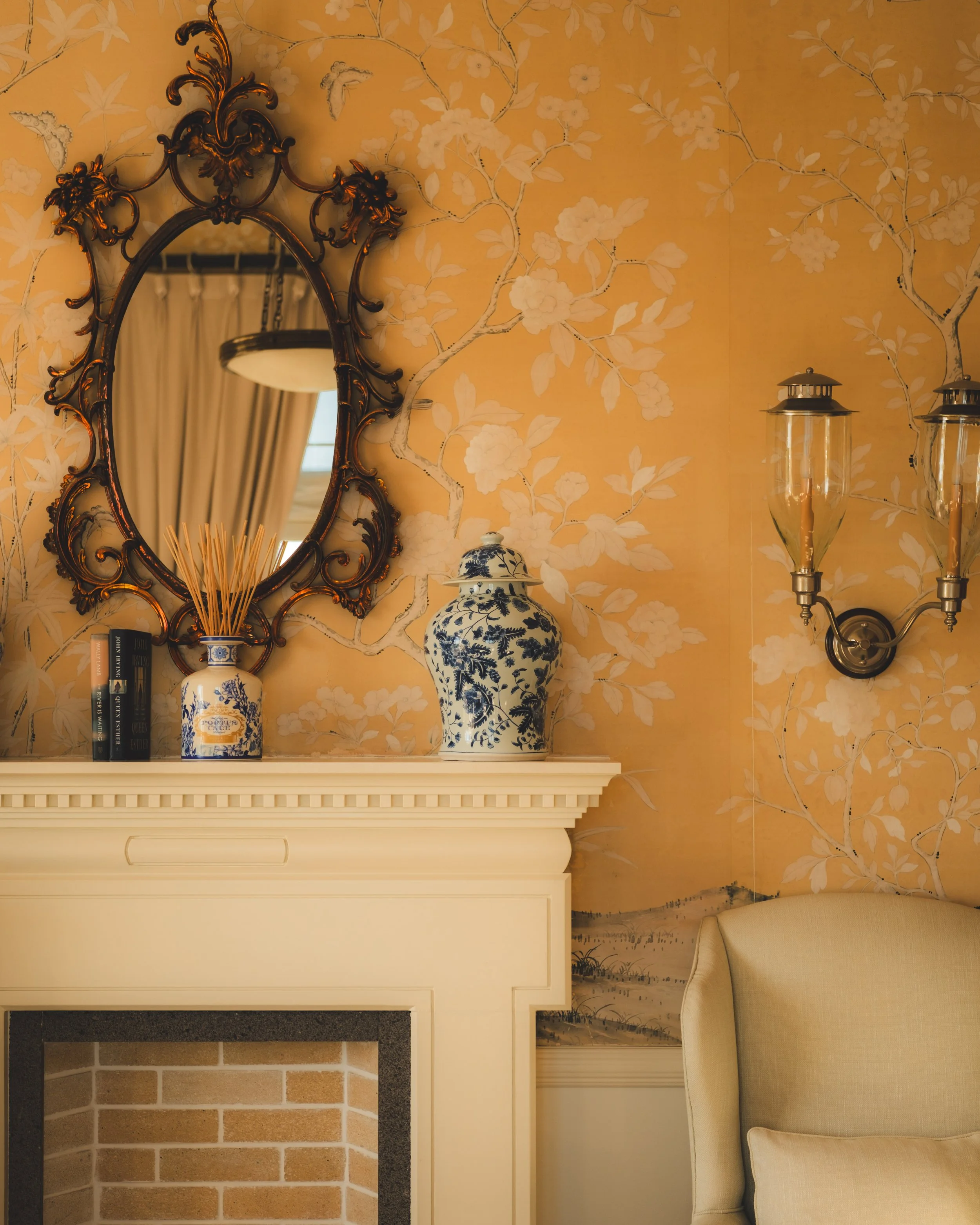 A living room corner with a beige armchair, a decorative fireplace mantle, a blue and white porcelain vase, and a wall mirror with an ornate frame. The wall has a floral wallpaper pattern with branches and flowers. There are two wall-mounted lanterns