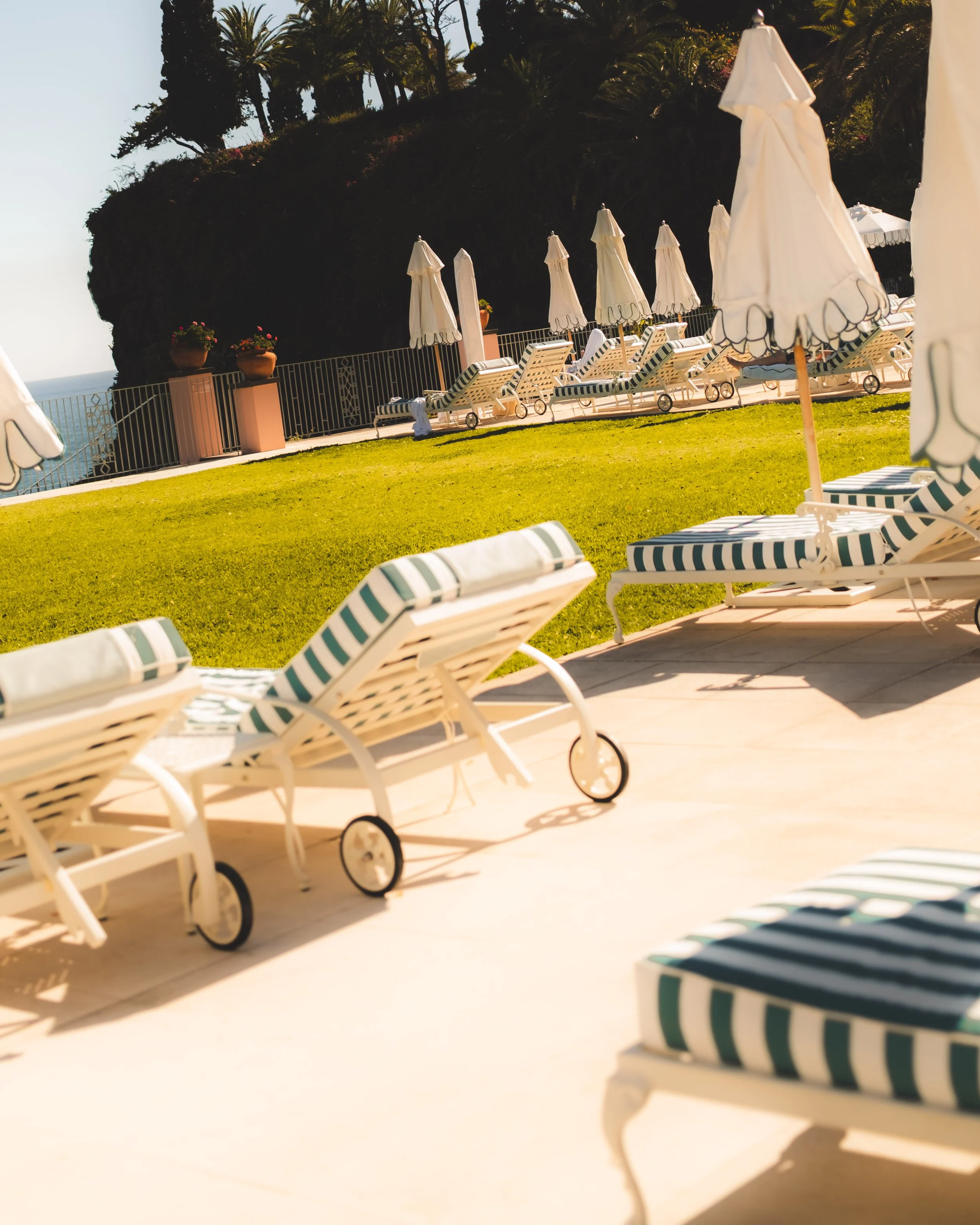 Empty lounge chairs and umbrellas on a grassy area by a fence overlooking the ocean, with a large rocky hill and trees in the background, under clear sunny sky.