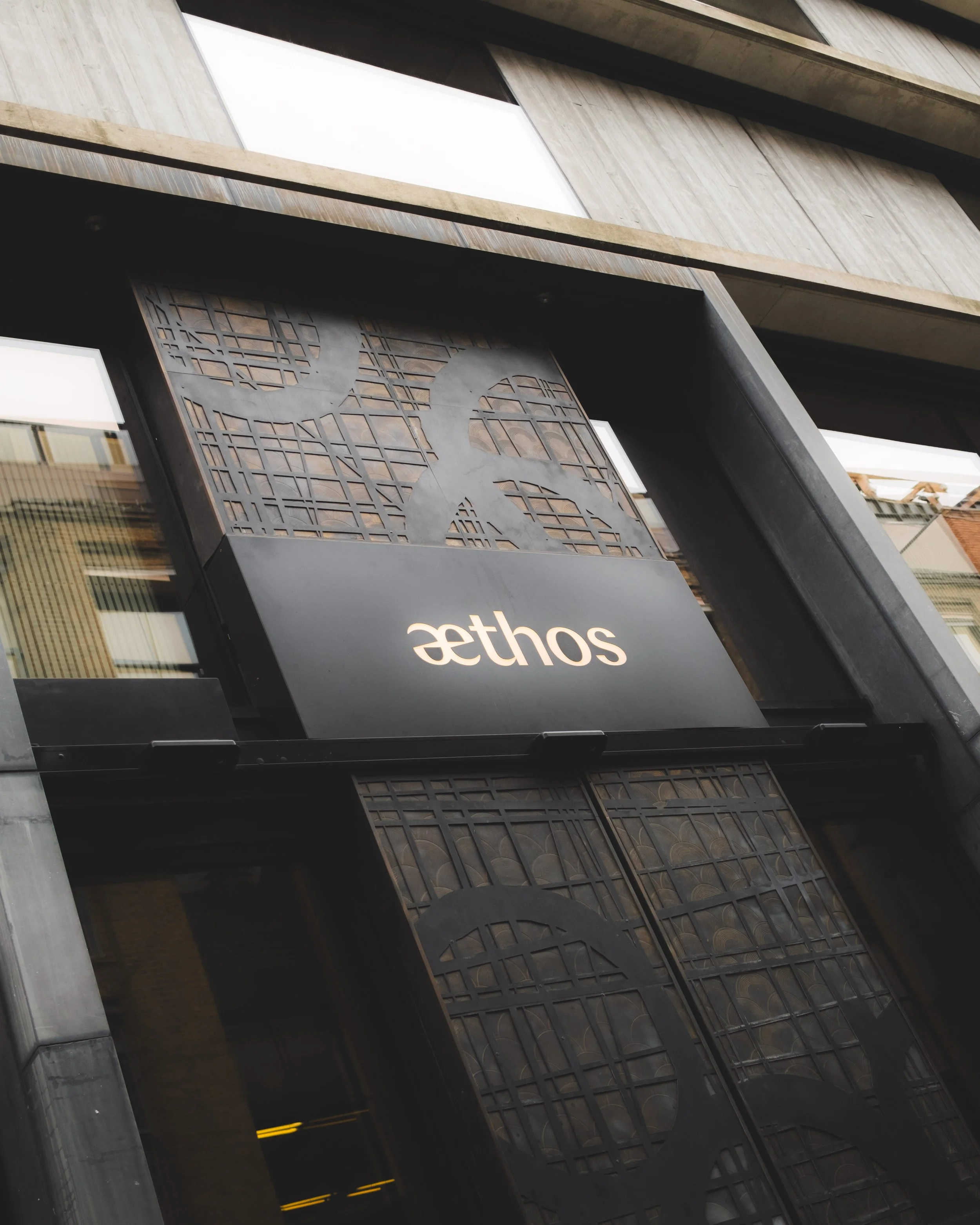 A modern building facade with concrete, glass, and decorative metal paneling, featuring a sign that reads 'Aethos'.